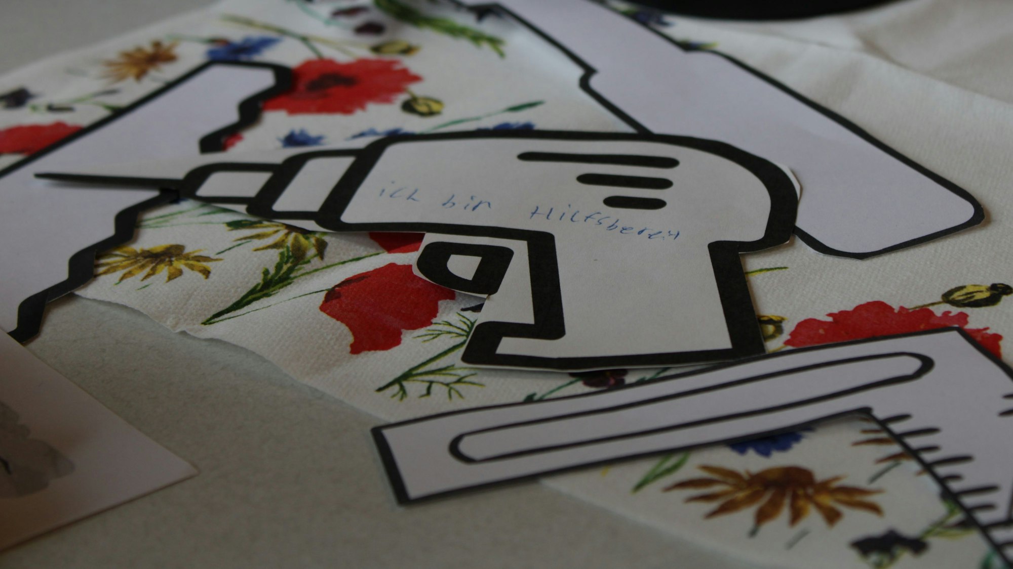 Verschiedene Papierschnipsel liegen auf einem Tisch, einer ist geformt wie eine Bohrmaschine, auf der steht: „Ich bin hilfsbereit“.