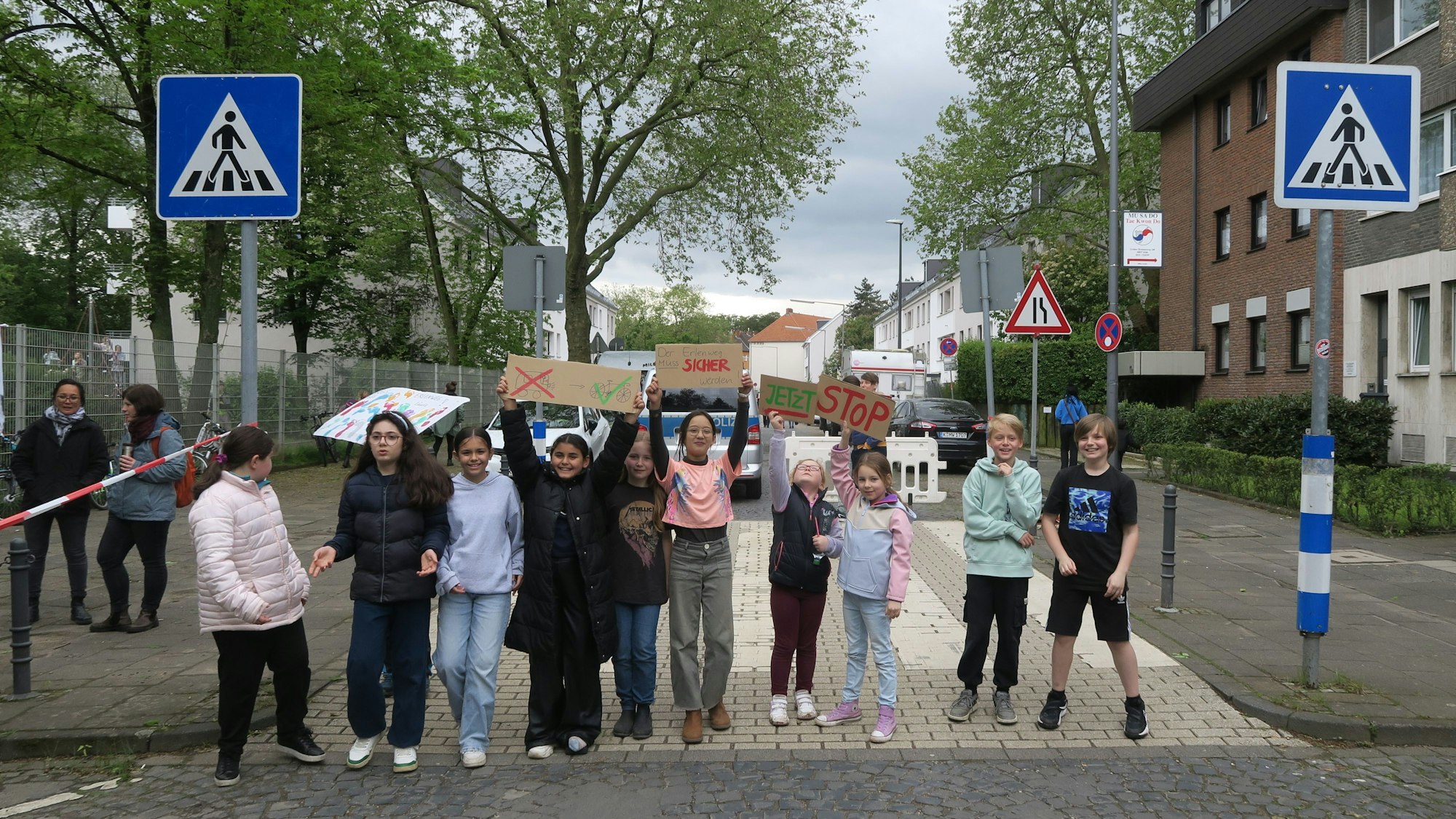 Mehrere Kinder stehen in einer Reihe und blockieren so eine Straße. Sie halten Plakate hoch, auf denen sie einen sicheren Schulweg fordern.
