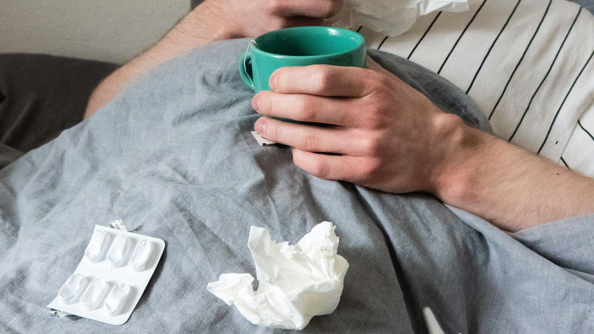 Ein Mann liegt mit Taschentüchern, Teetasse, Nasenspray und Tabletten im Bett (gestellte Szene).