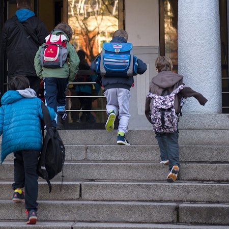 Grundschulkinder laufen mit ihren Ranzen auf dem Rücken eine Treppe hoch zum Schuleingang.