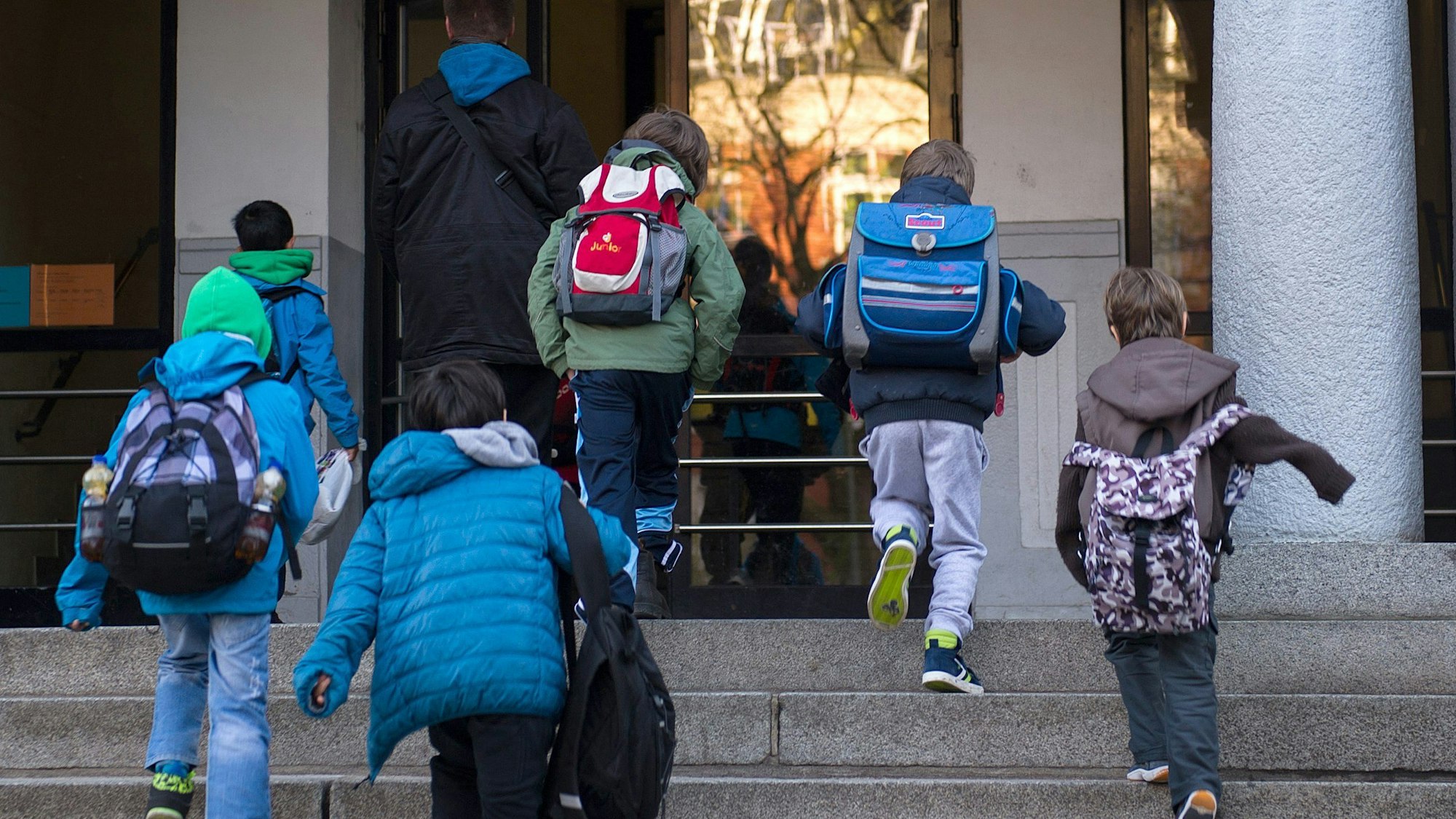 Grundschulkinder laufen mit ihren Ranzen auf dem Rücken eine Treppe hoch zum Schuleingang.