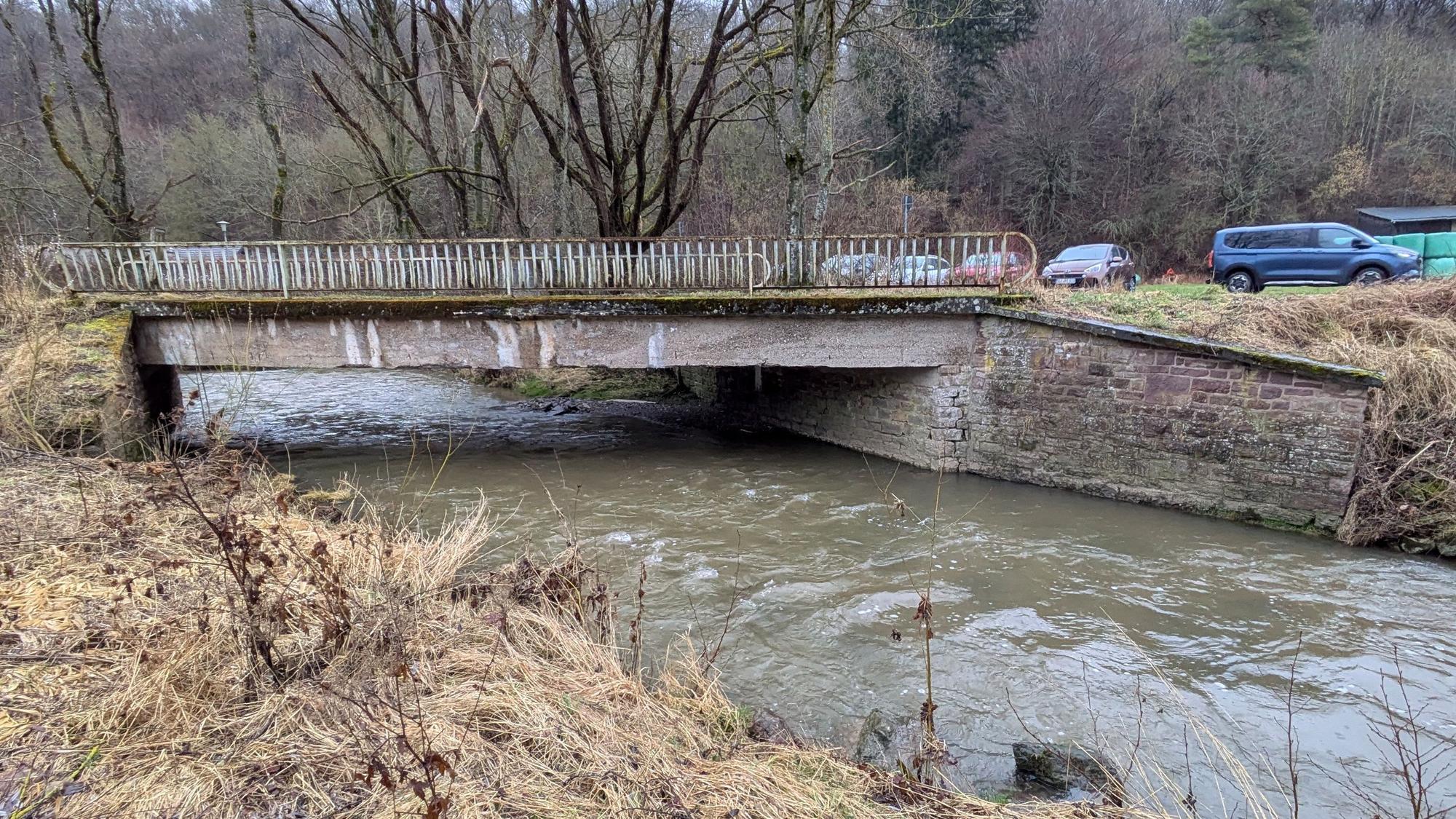 Eine Wirtschaftswegebrücke führt bei Nettersheim über die Urft. Im Hintergrund parken mehrere Autos.
