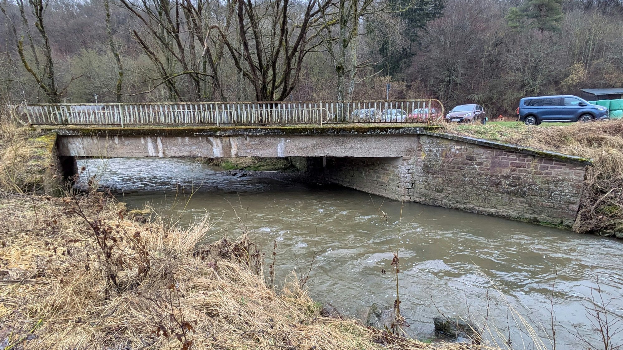 Eine Wirtschaftswegebrücke führt bei Nettersheim über die Urft. Im Hintergrund parken mehrere Autos.