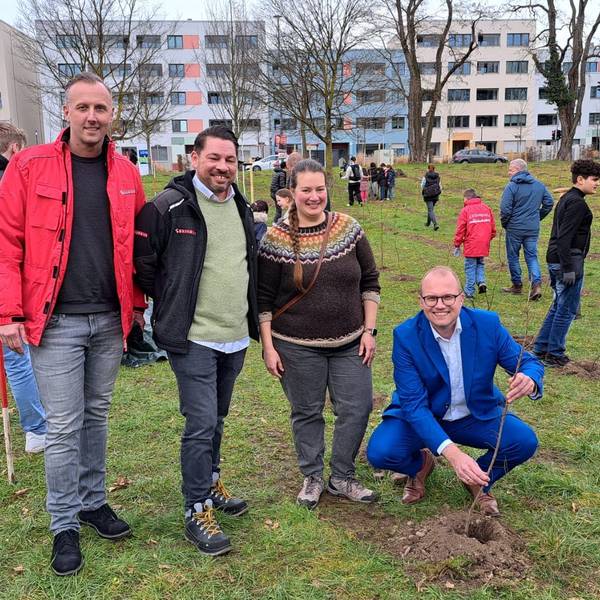 Drei Männer und eine Frau stehen und hocken auf einer Wiese, um sie herum sind viele Kinder. Ein Mann hält einen jungen Baum, der gerade gepflanzt wurde.