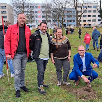 Drei Männer und eine Frau stehen und hocken auf einer Wiese, um sie herum sind viele Kinder. Ein Mann hält einen jungen Baum, der gerade gepflanzt wurde.
