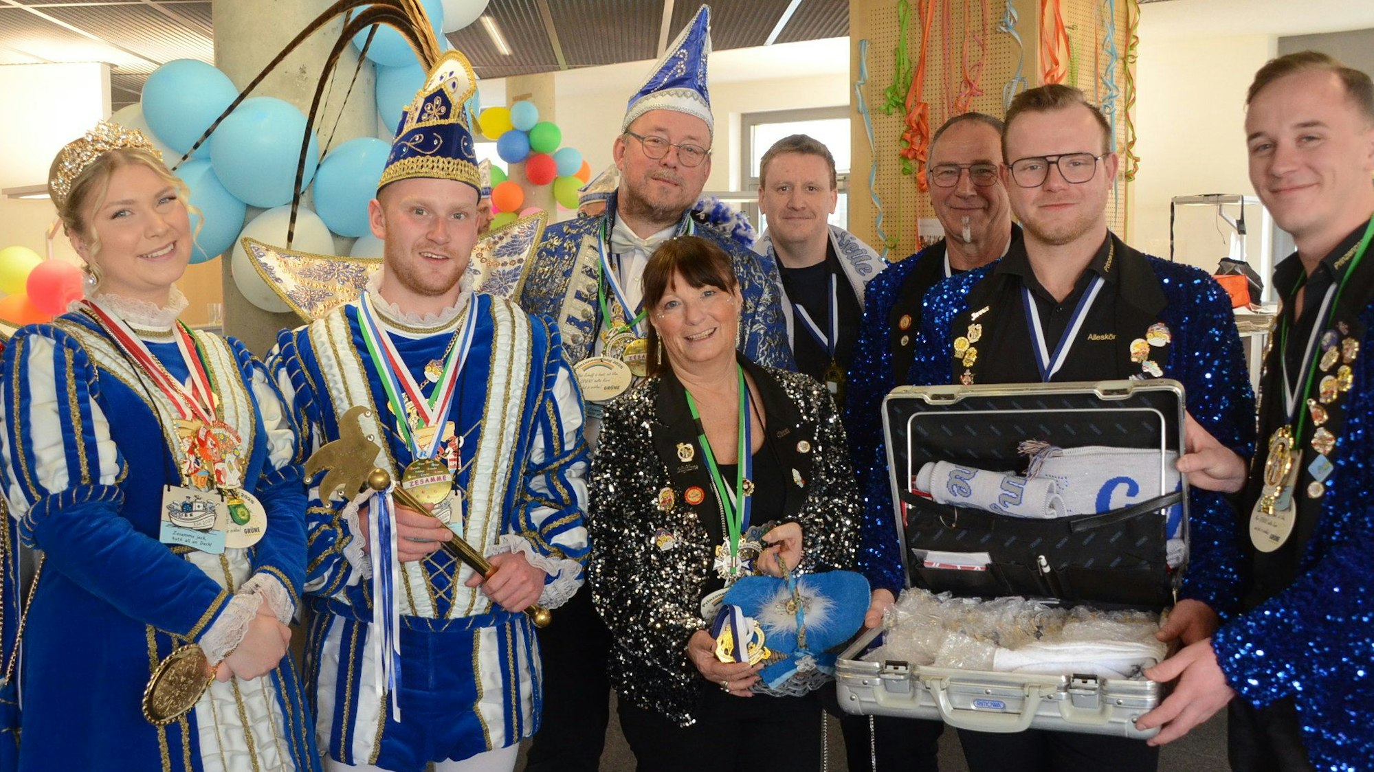 Ein Karnevalsprinzenpaar und seine Begleitung. Zwei Männer zeigen den Inhalt eines geöffneten Koffers.