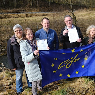 Die Genannten stehen vor einer Brachfläche an der Urft. Sie halten eine EU-Fahne und Vertragsunterlagen hoch.