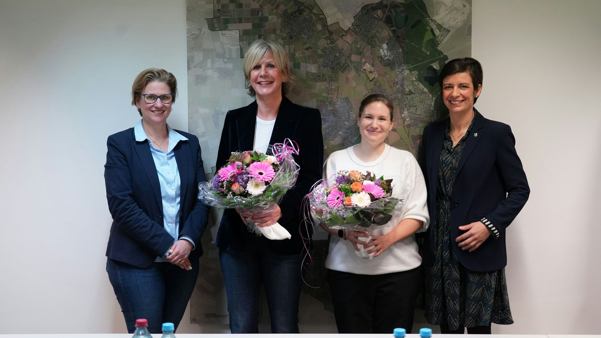 Vier Frauen lächeln in die Kamera, die beiden in der Mitte halten Blumensträuße in ihren Händen.