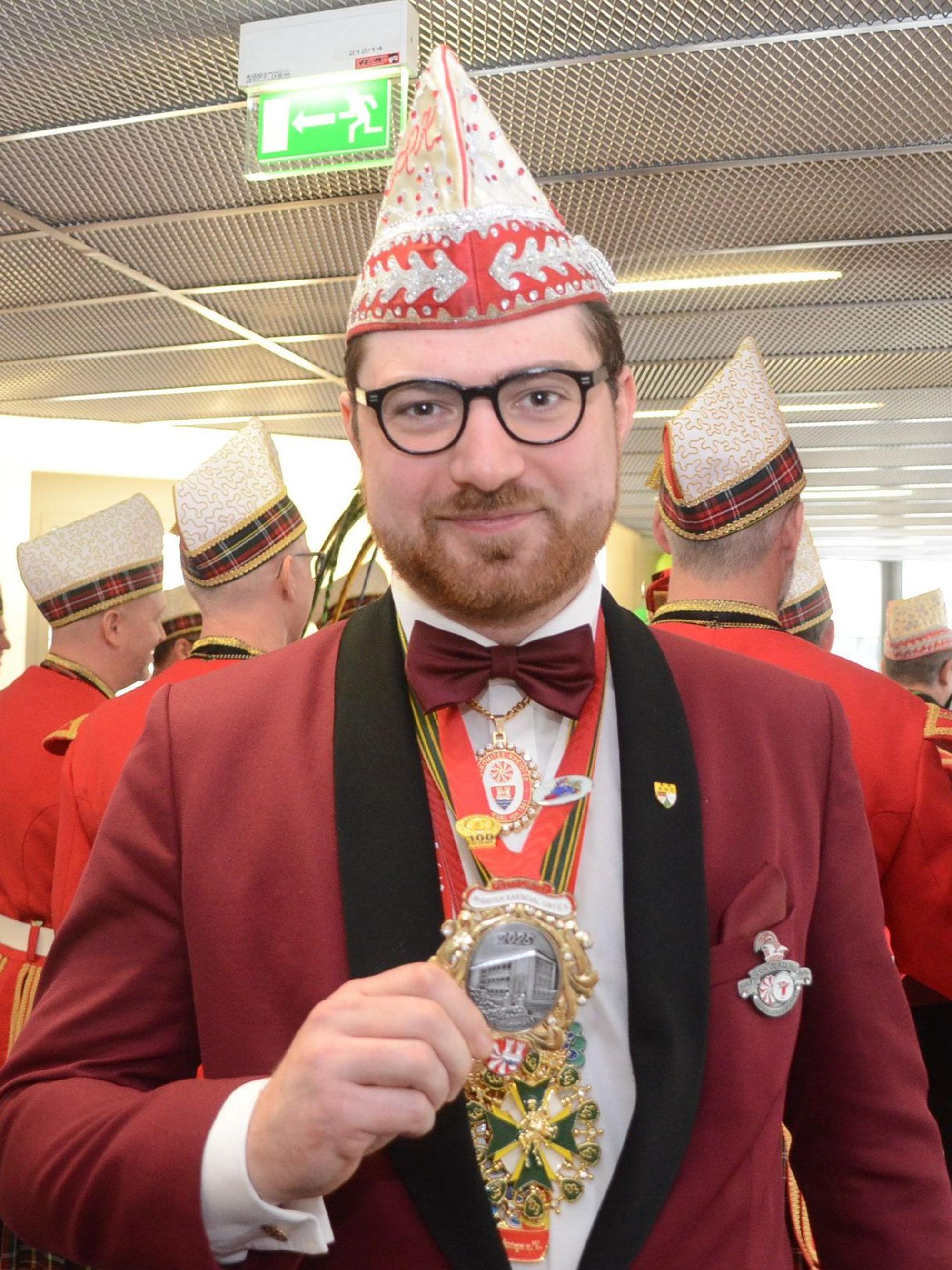 Ein Mann mit dem Schiffchen einer Karnevalsgesellschaft, in dunkelroter Jacke mit schwarzen Revers. Er hält einen Orden in die Kamera.