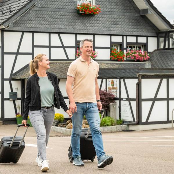 Zwei Menschen ziehen einen Rollkoffer hinter sich her. Im Hintergrund ist ein Fachwerkhaus zu sehen.