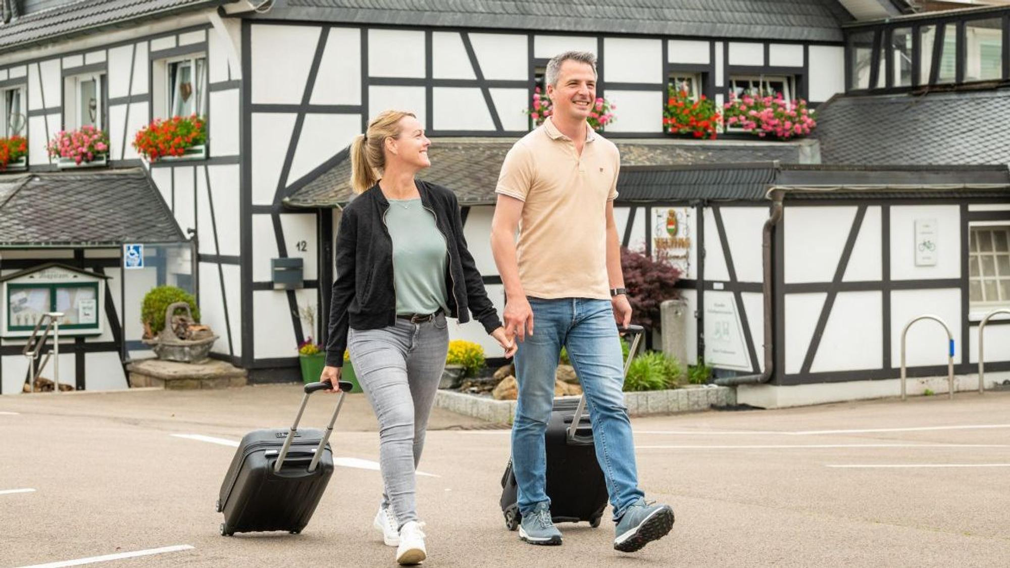 Zwei Menschen ziehen einen Rollkoffer hinter sich her. Im Hintergrund ist ein Fachwerkhaus zu sehen.