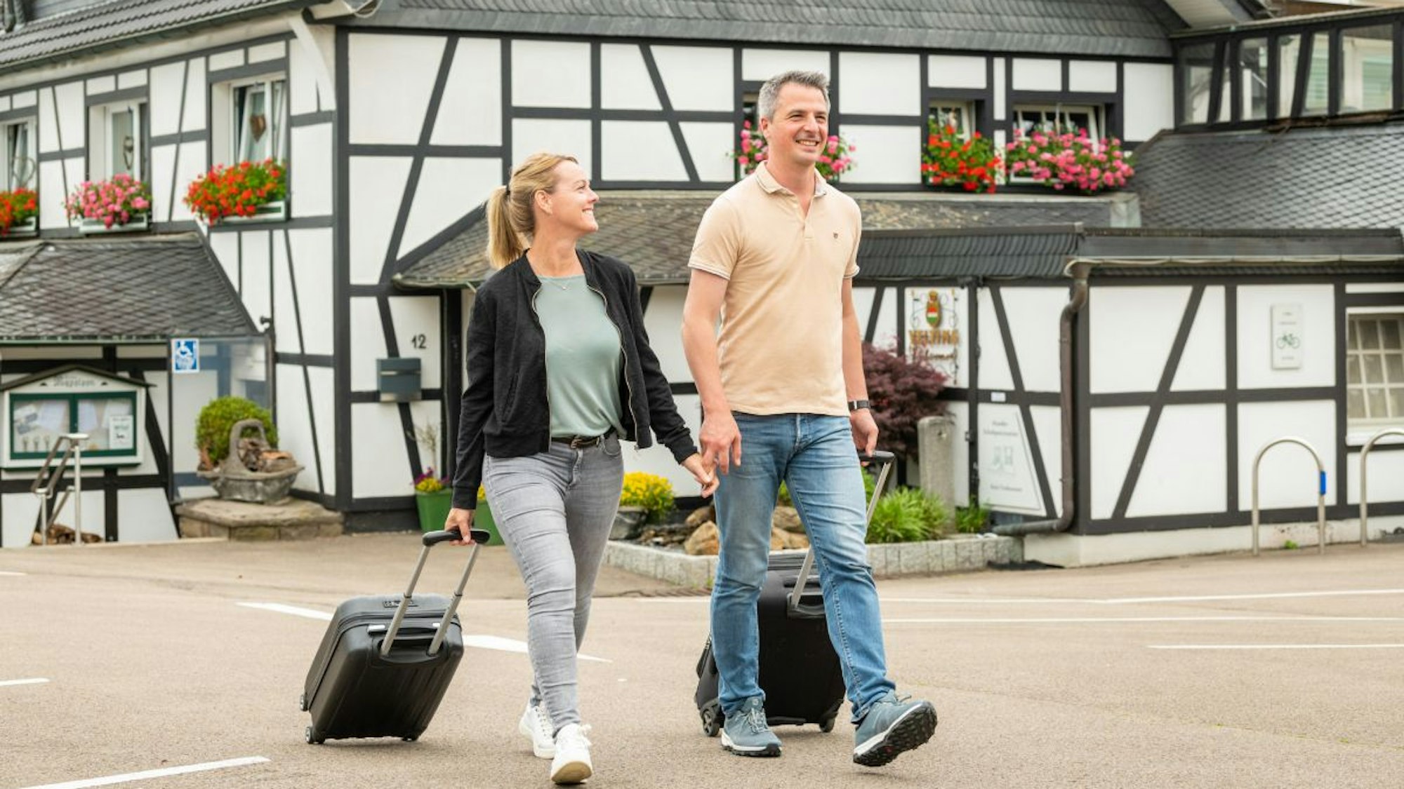 Zwei Menschen ziehen einen Rollkoffer hinter sich her. Im Hintergrund ist ein Fachwerkhaus zu sehen.