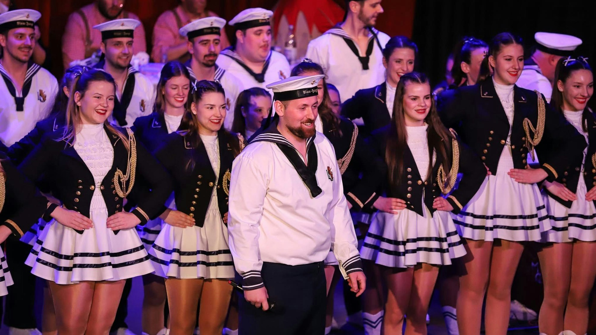 Auf dem Bild ist ein Mann im Matrosenkostüm vor einer Reihe Tänzer auf der Bühne zu sehen.