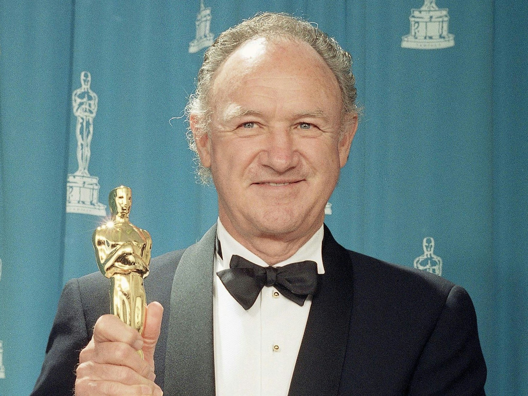 Gene Hackman hält einen Oscar in der Hand und lächelt.