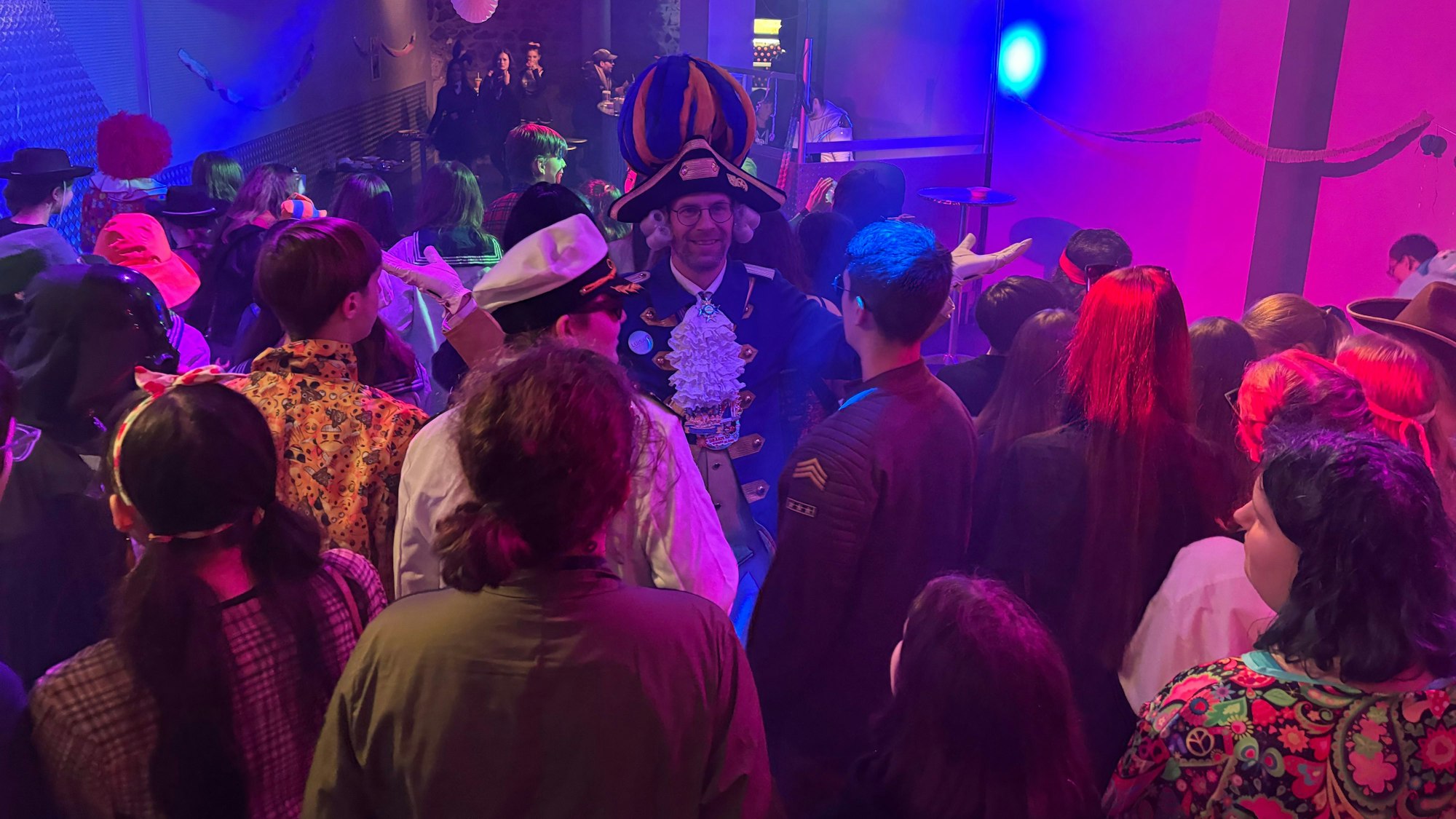 Ein kostümierter Mann steht inmitten von Jugendlichen, die von der Kamera wegschauen. Er streckt die Arme mit nach oben gedrehten Handflächen von sich.