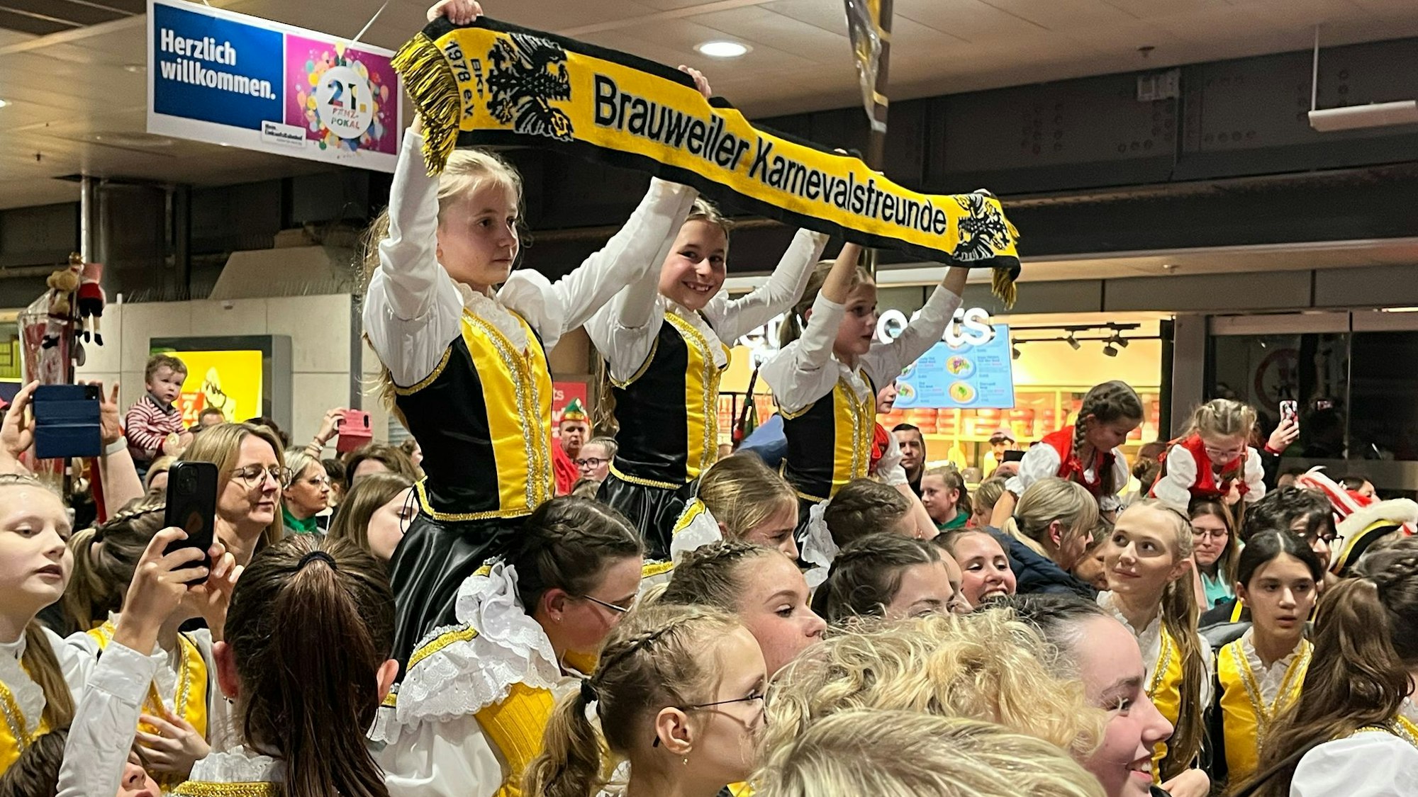 Die Pänz tragen gelb-schwarze Trachten. Drei von ihnen werden auf den Schultern ihrer Mittäänzerinnen getragen und halten einen Schal hoch, auf dem „Brauweiler Karnevalsfreunde“ steht.