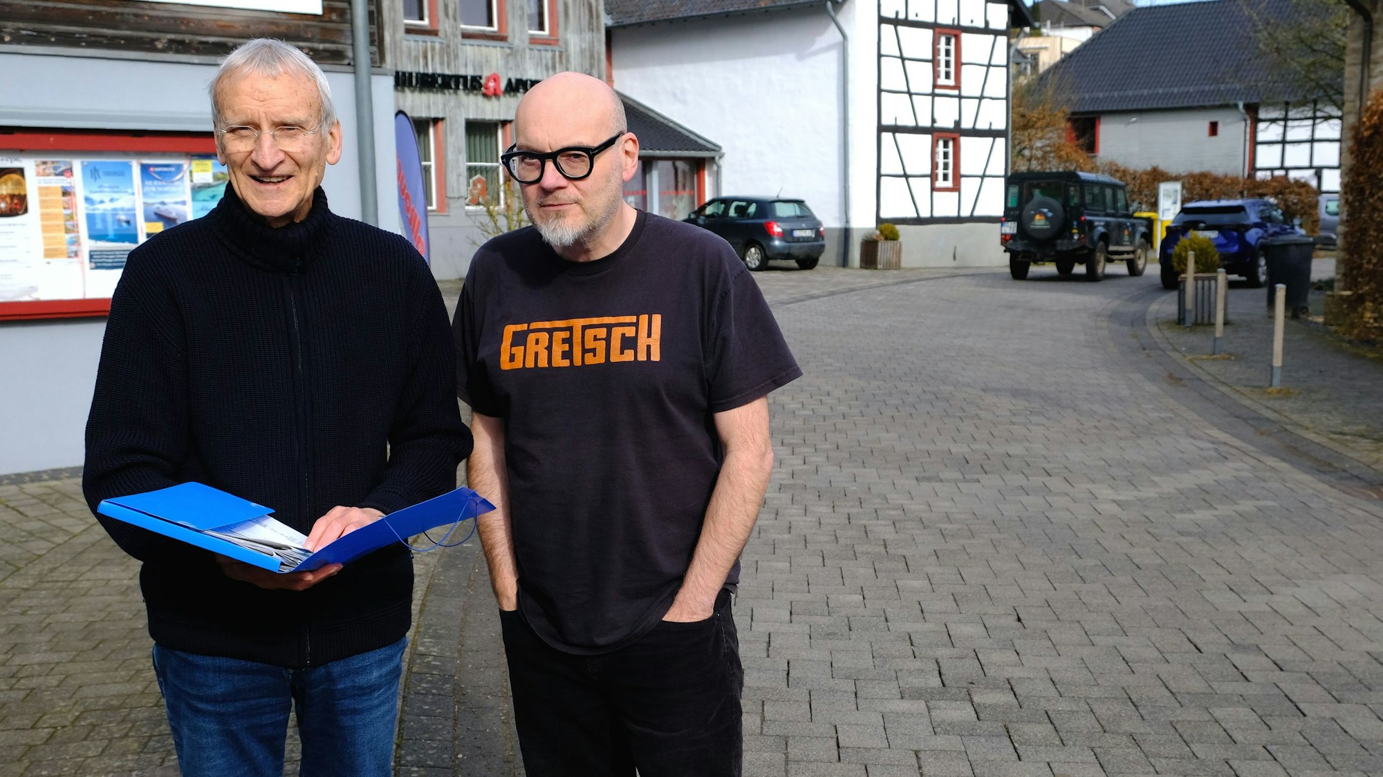 Franz-Josef Hilger und Andreas Reck stehen auf der Steinfelder Straße. Hilger hält eine blaue Mappe mit Unterlagen in den Händen, Reck hat die Hände in den Hosentaschen. Im Hintergrund fährt ein Jeep über die Straße.