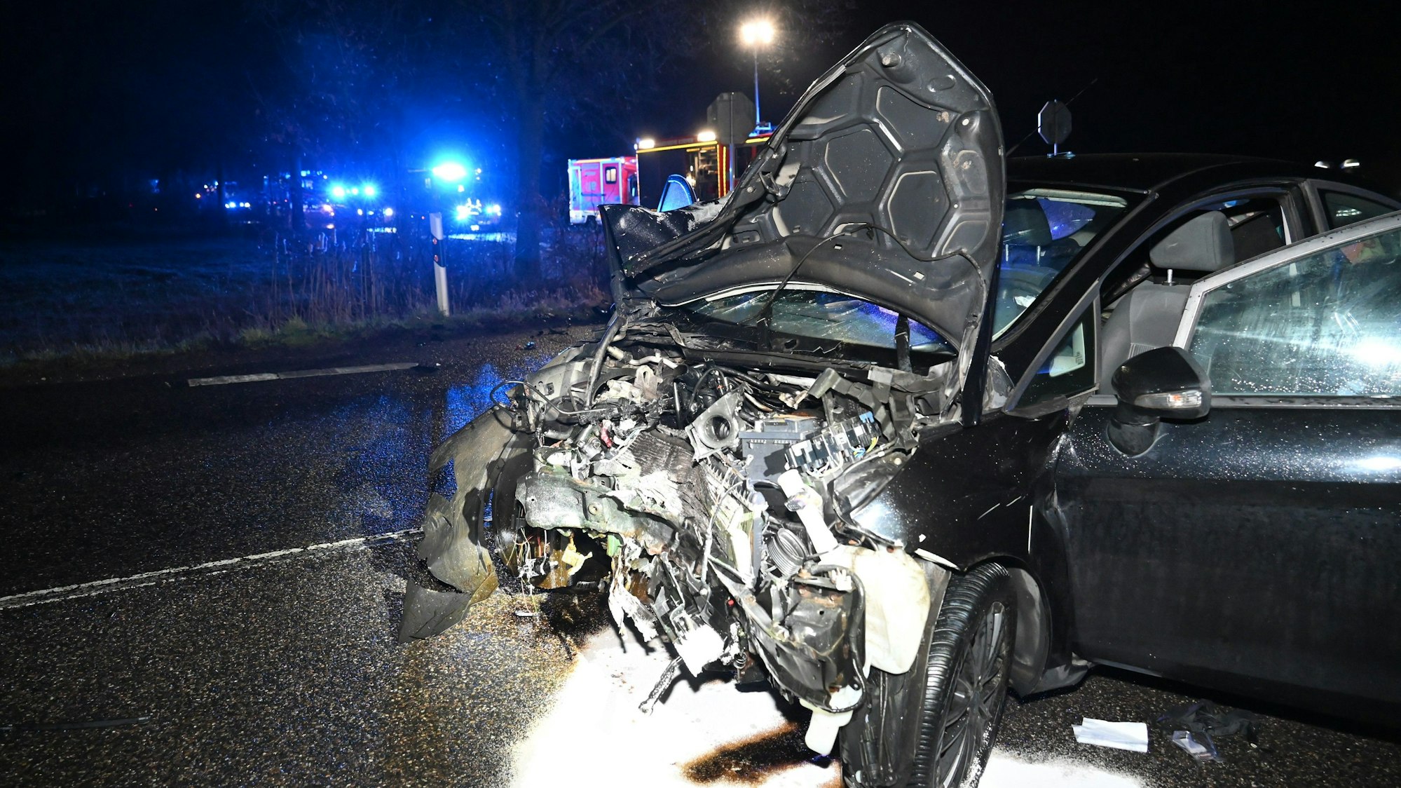 Ein dunkles Auto mit komplett zerstörter Motorhaube steht auf einer Straße. Im Hintergrund ist Blaulicht zu sehen.
