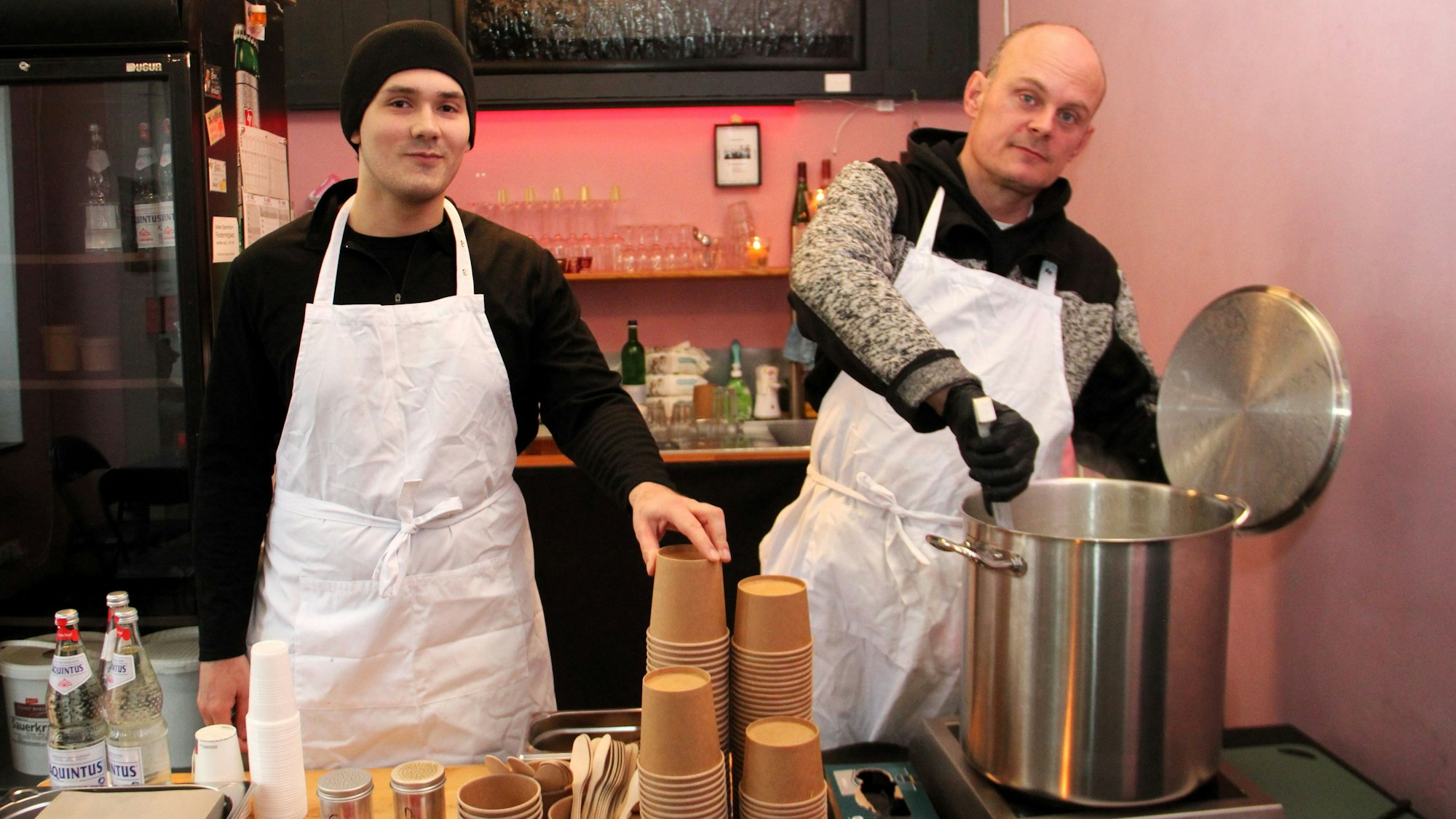 Zwei Männer mit weißen Schürzen stehen hinter einem Tisch. Einer von ihnen rührt mit einer Suppenkelle in einem großen Kochtopf.