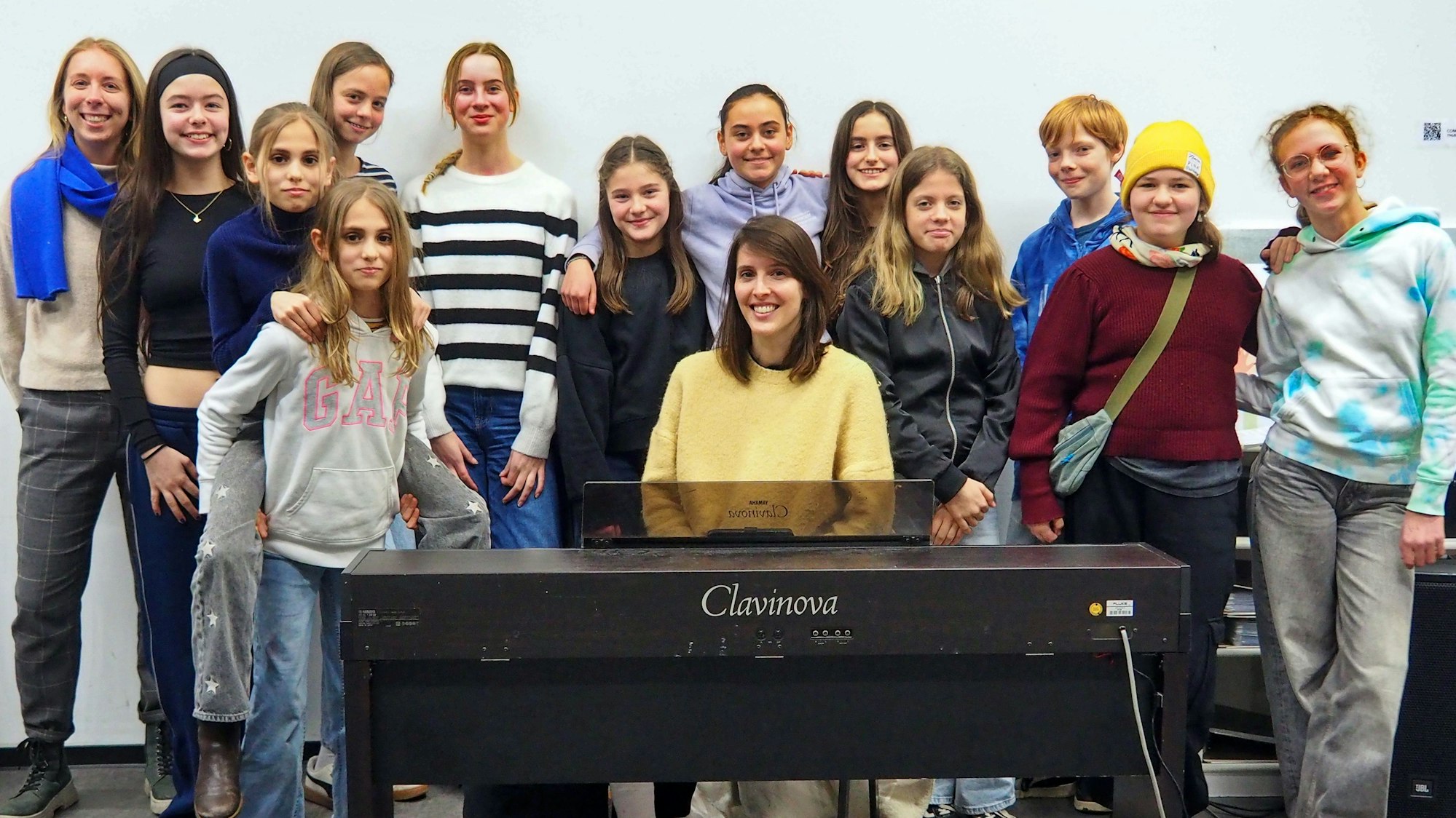 Um ein Klavier herum stehen etwa ein Dutzend Jugendliche. Die Leiterin des Chores sitzt am Klavier.