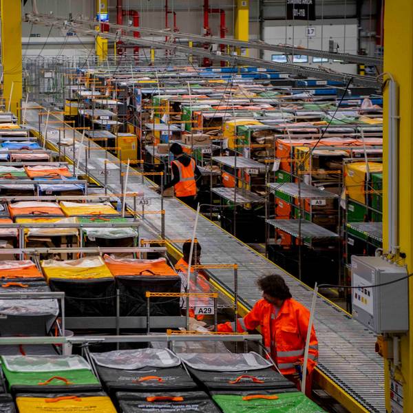 In einer Halle des Amazon-Verteilzentrums im IPAS in Euskirchen sind mehrere Mitarbeiter in orangefarbenen Westen und Jacken sowie Reihen mit verschiedenfarbigen Taschen zu sehen.