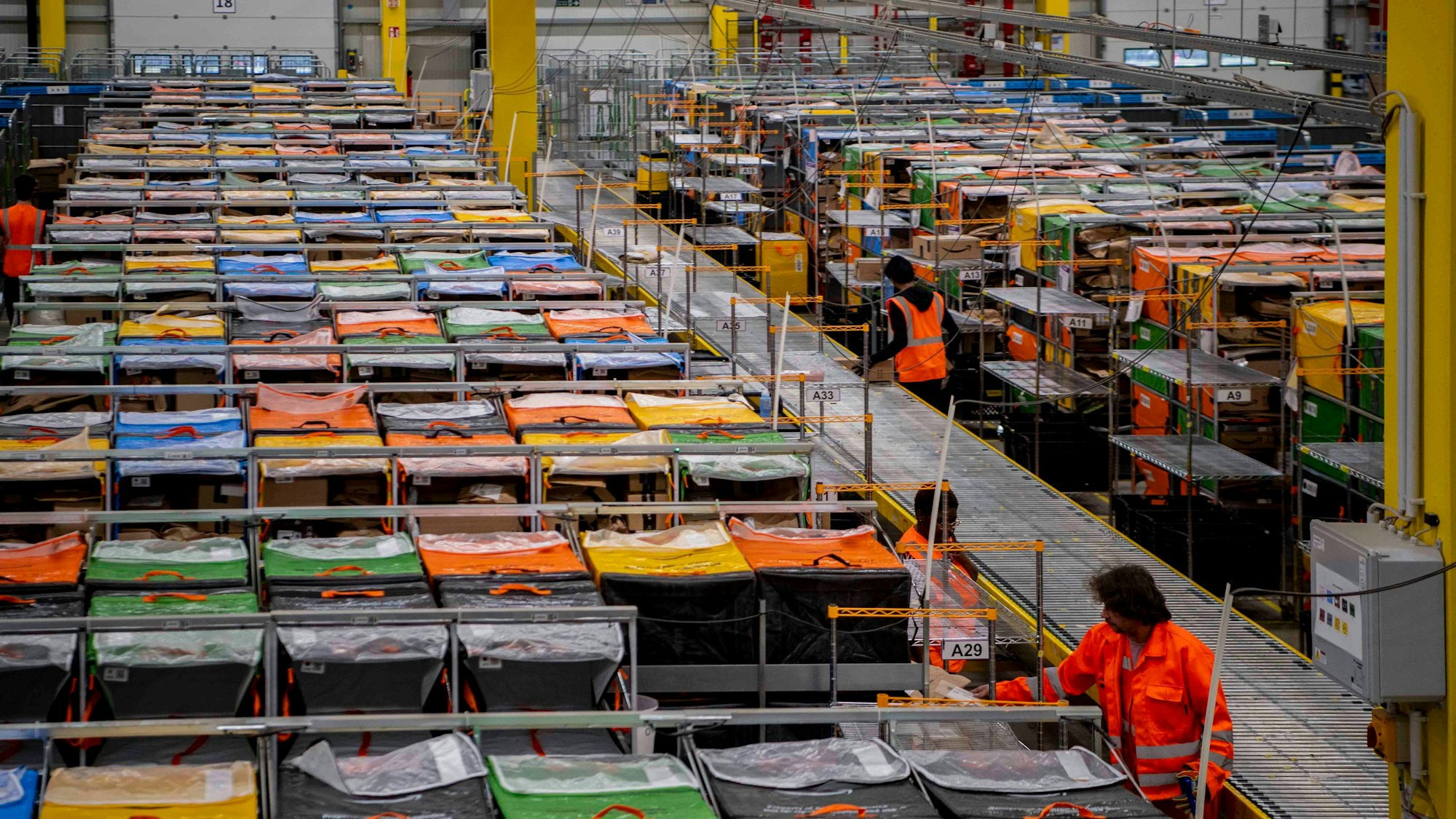 In einer Halle des Amazon-Verteilzentrums im IPAS in Euskirchen sind mehrere Mitarbeiter in orangefarbenen Westen und Jacken sowie Reihen mit verschiedenfarbigen Taschen zu sehen.
