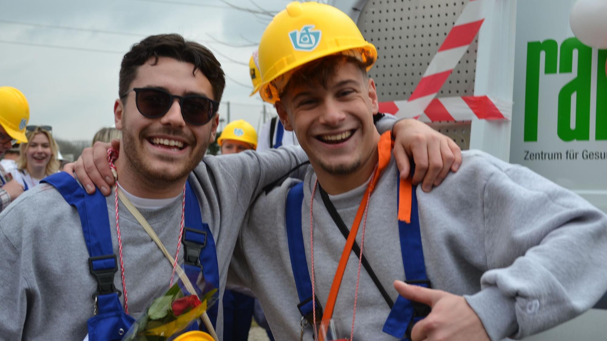 Zwei lachende Männer in Latzhose und mit Bauhelmen.
