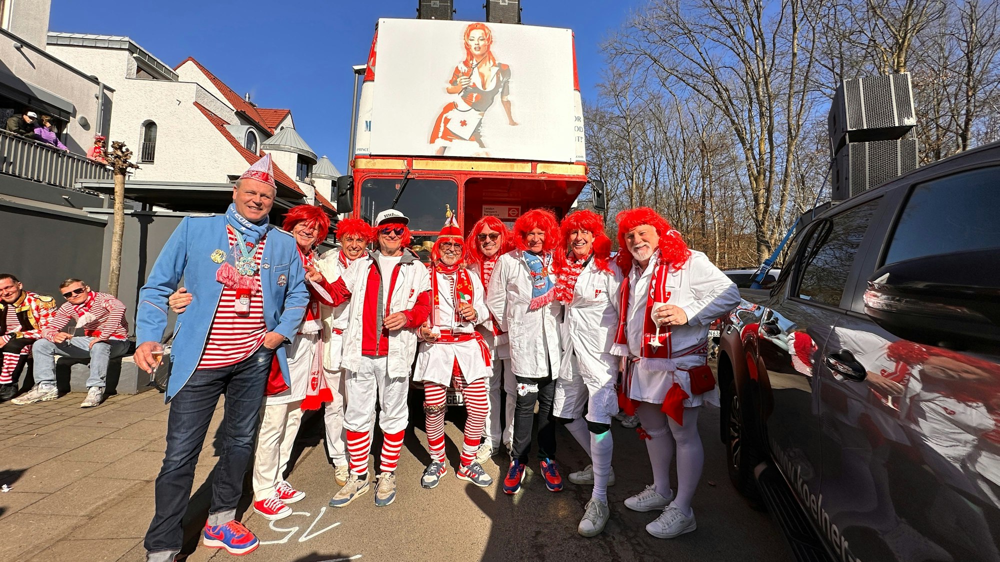 Eine Grupppe Männer in Krankenschwester-Kostümen mit roten Perücken steht vor einem Doppeldecker-Bus.