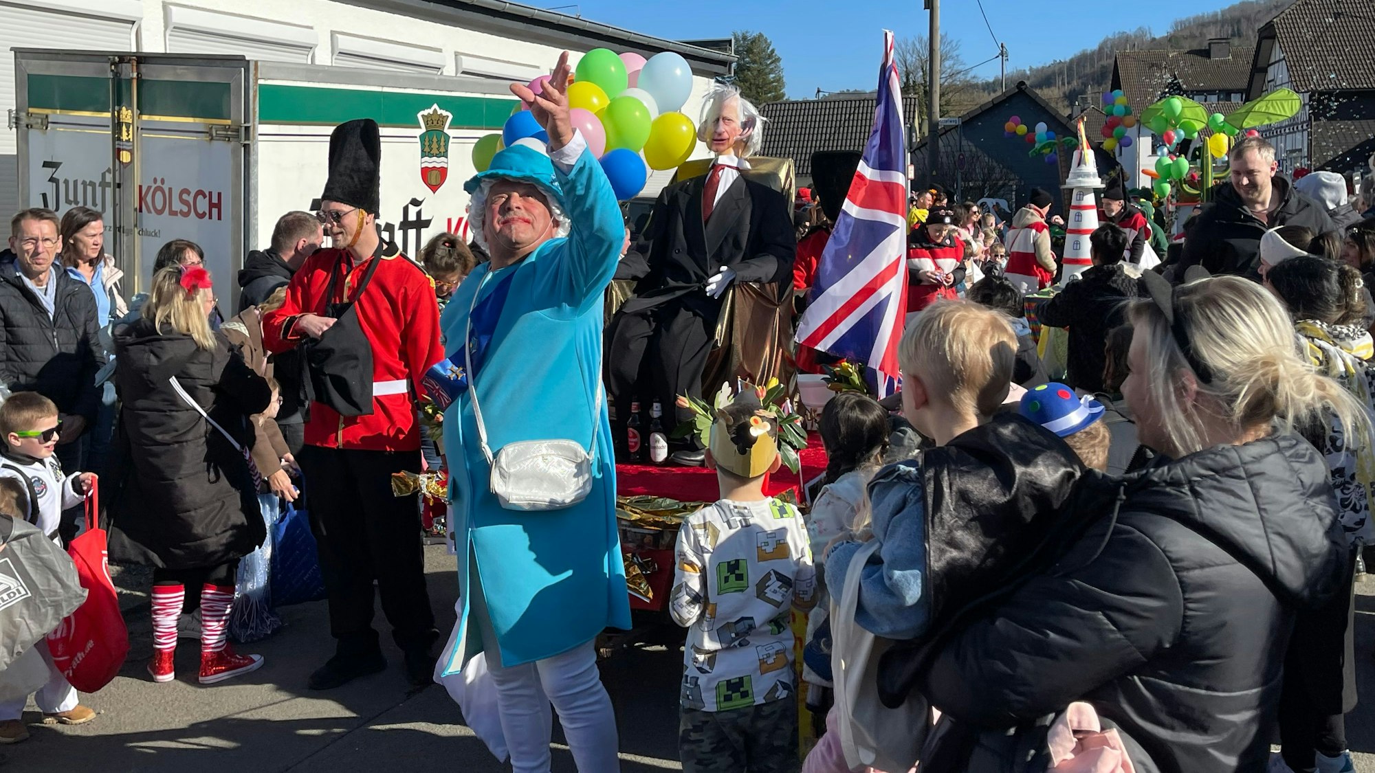 Ein Mann bei einem Karnevalszug, der als Queen Elizabeth II. verkleidet ist, wirft Kamelle in die Menge.