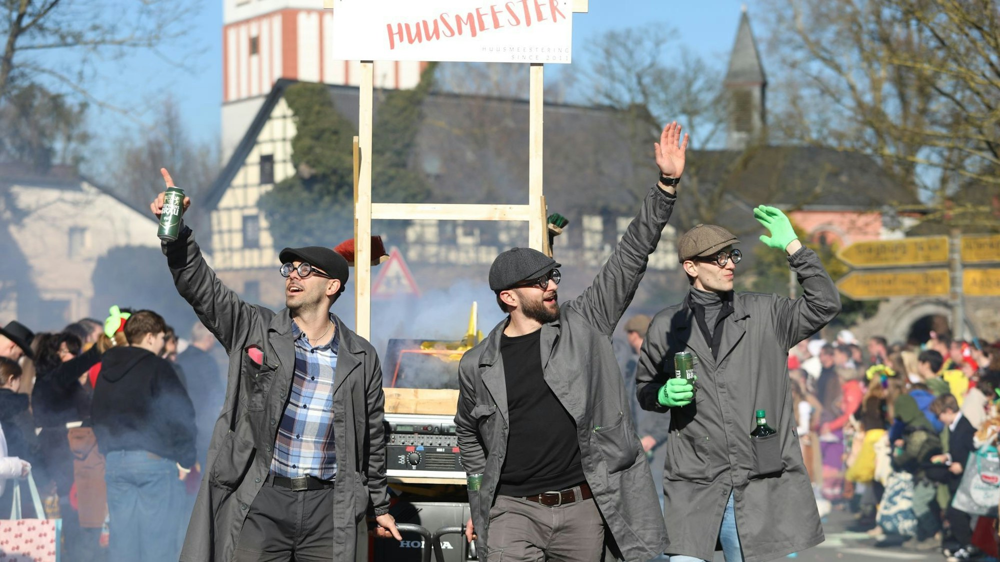 Drei Männer in Hausmeisterkleidung ziehen einen Karnevalswagen, im Hintergrund die Oberpleiser Pfarrkirche.