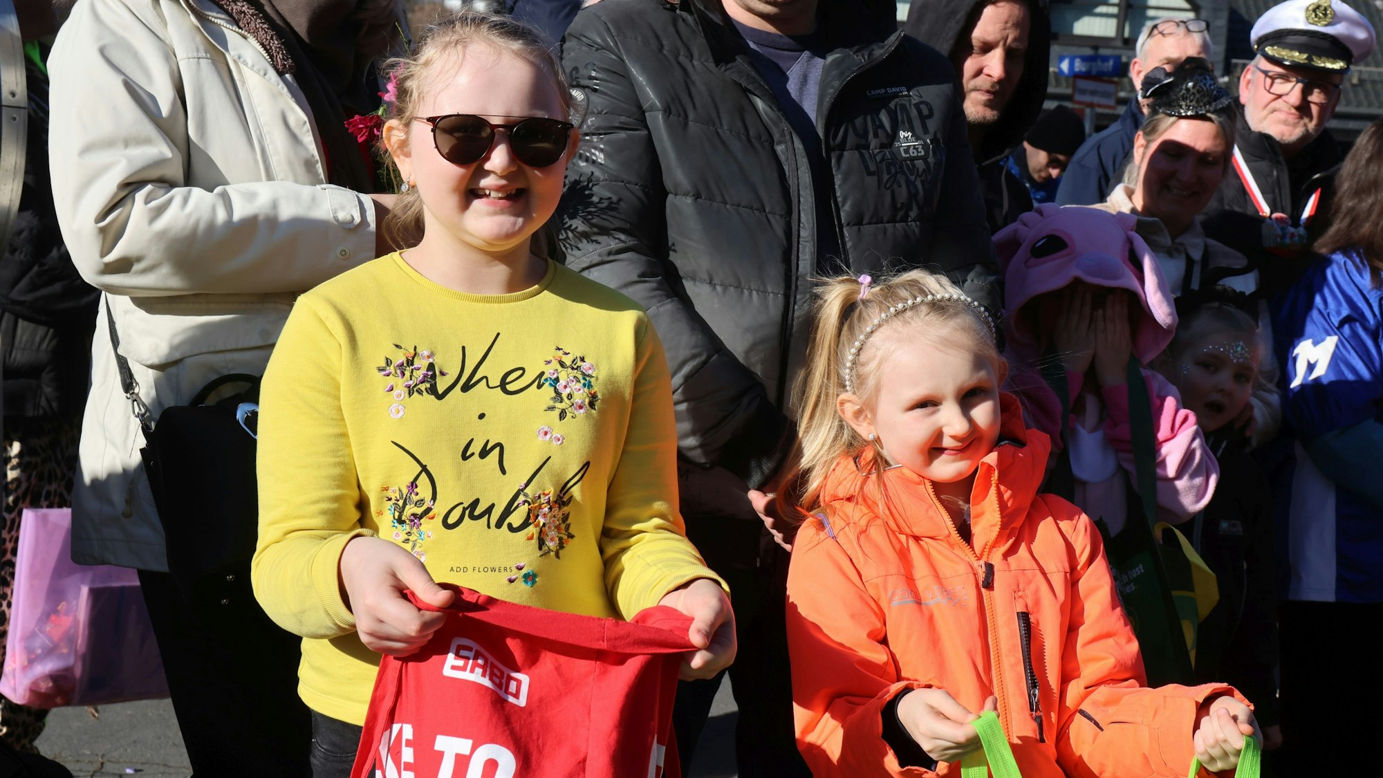 Zwei Kinder stehen mit geöffneten Tüten am Rande eines Karnevalszugs.
