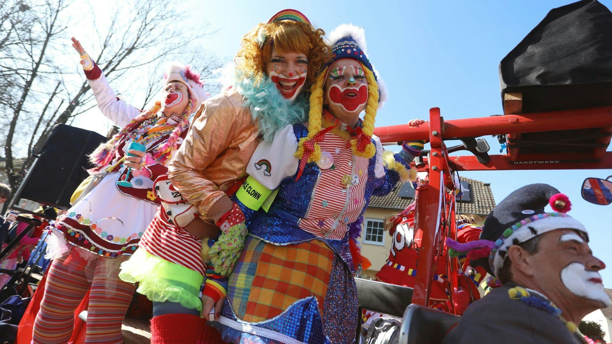 Drei bunt kostümierte und geschminkte Frauen als Clowns auf einem Karnevalswagen.