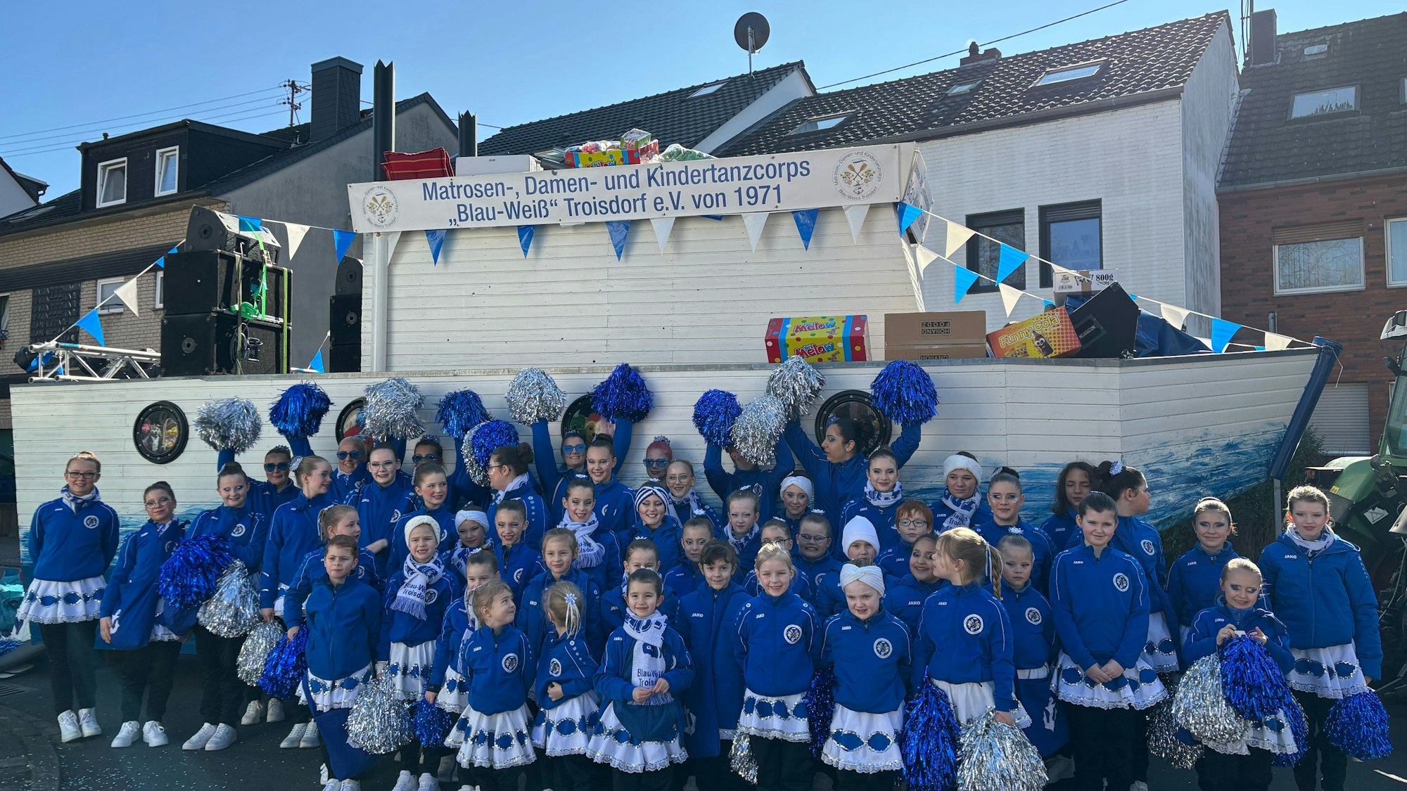 Eine Gruppe mit Kindern in blau-weißen Kostümen steht vor einem Karnevalswagen, der wie ein Schiff aussieht.