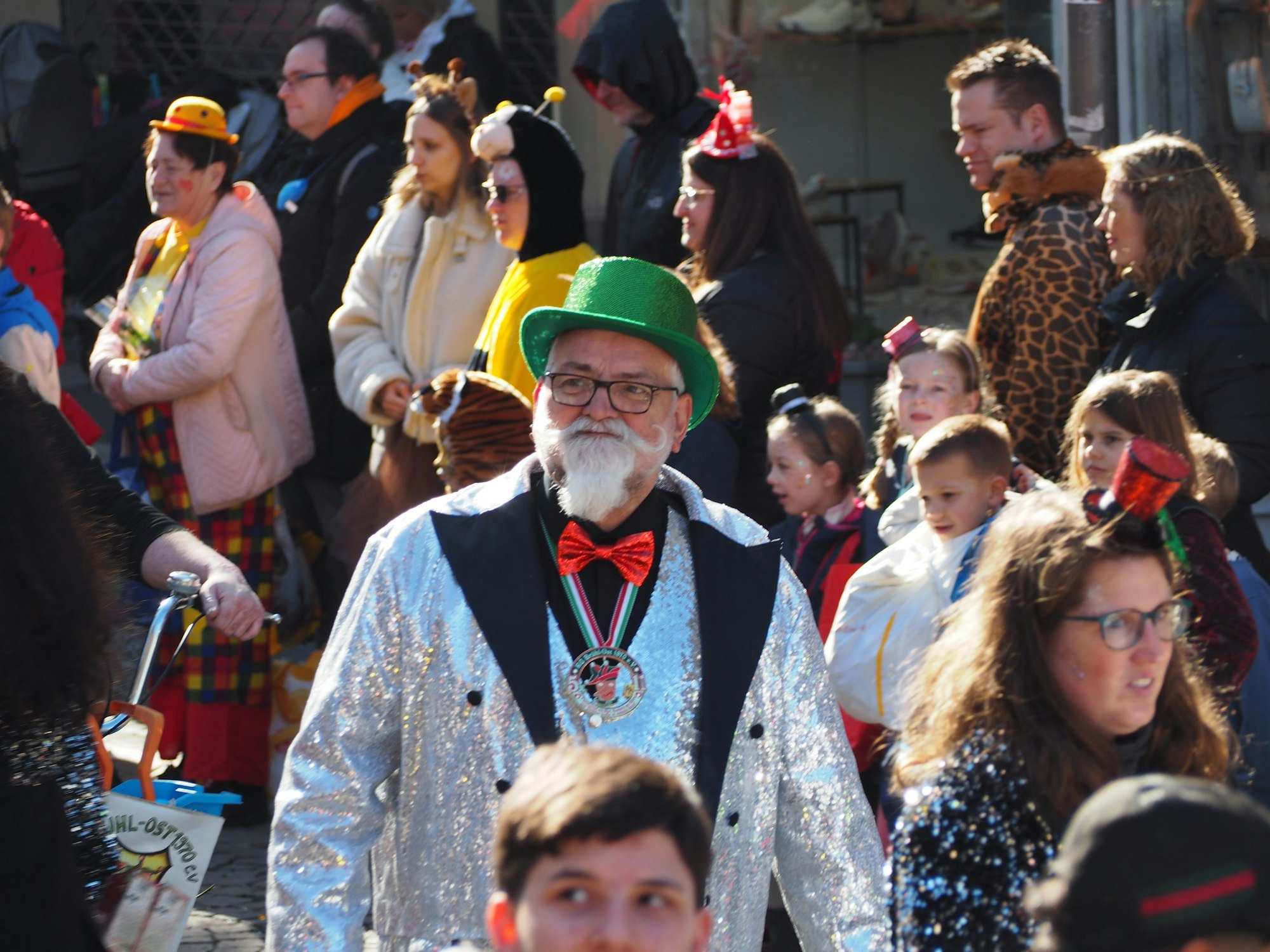 Ein Mann mit einem weißen Bart. Er trägt eine silbern-glitzernde Jacke und einen grünen Hut.