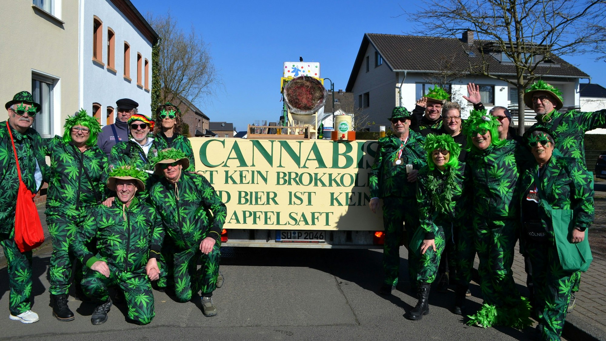 Menschen, die sich als Cannabis-Pflanzen verkleidet haben, flankieren einen Zug, auf dem eine große Joint-Installation mitfährt.