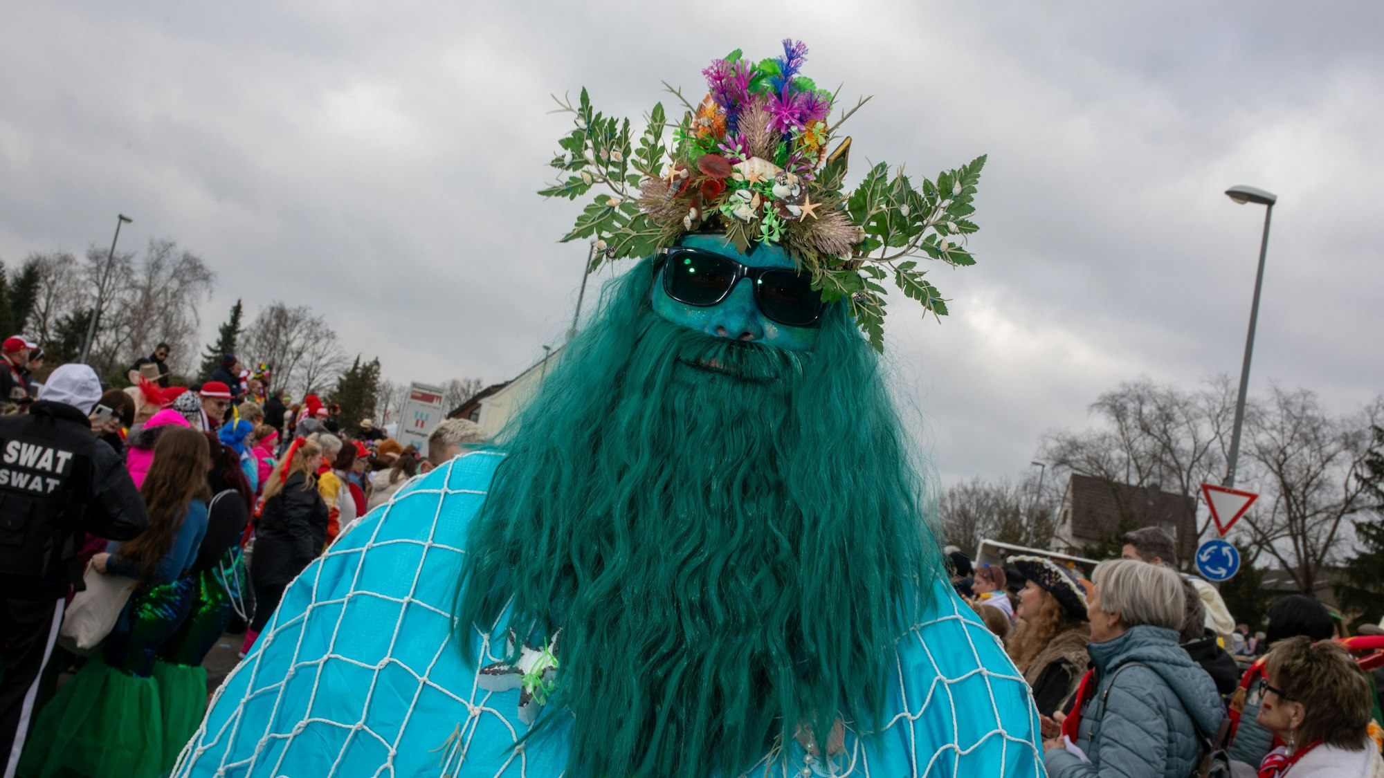Ein Mann mit riesigem grünen Bart, blauem Gesicht und einem Blumengesteck auf dem Kopf ist zu sehen.
