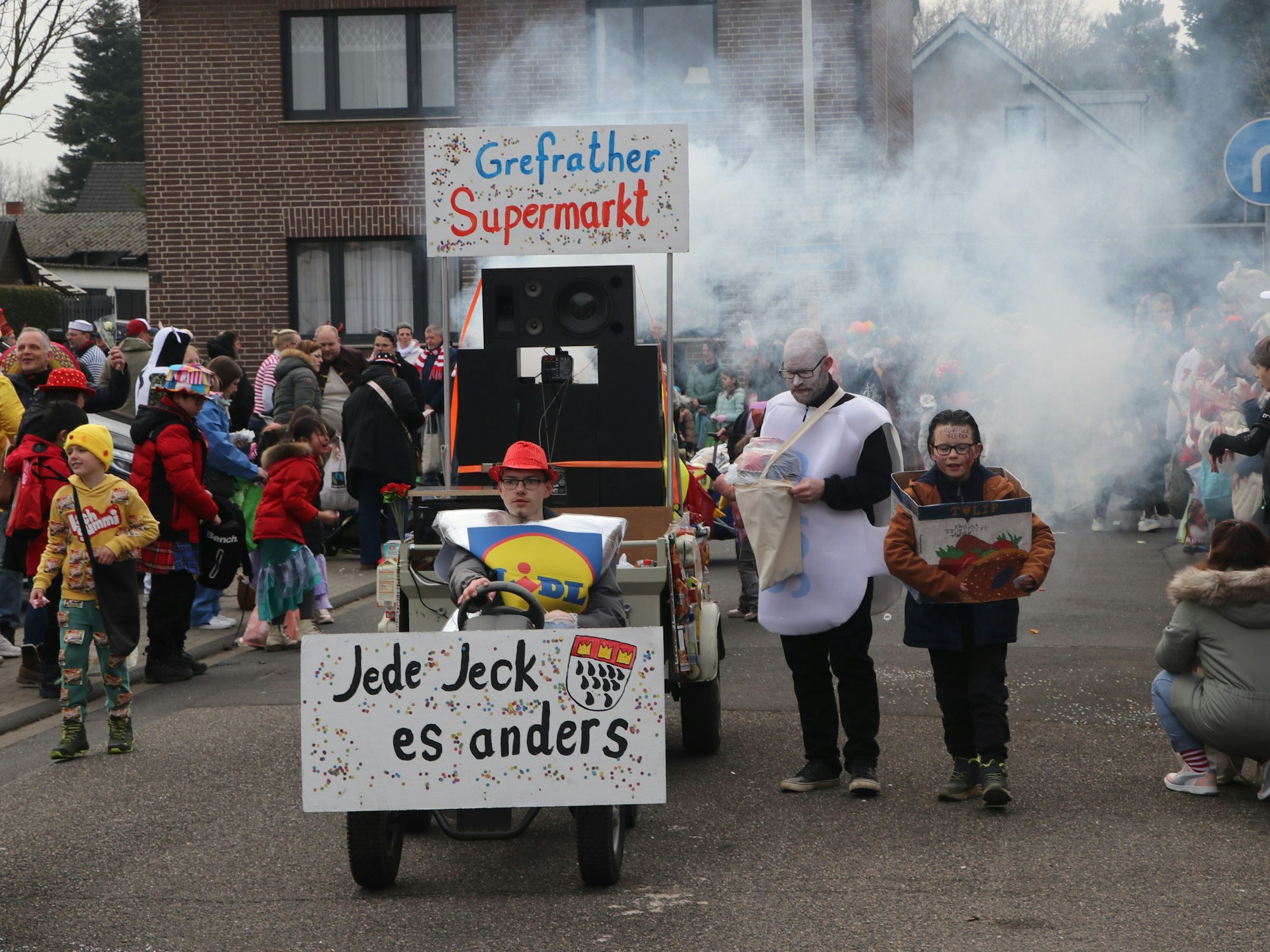 Auf dem Bild sind ein kleiner Karnevalswagen und Jecke am Straßenrand zu sehen.