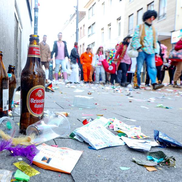 Zu sehen ist eine Ansammlung von Müll und leeren Bierflaschen auf einer Straße. Im Hintergrund sind Karnevalisten zu sehen.