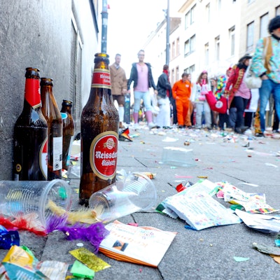 Zu sehen ist eine Ansammlung von Müll und leeren Bierflaschen auf einer Straße. Im Hintergrund sind Karnevalisten zu sehen.