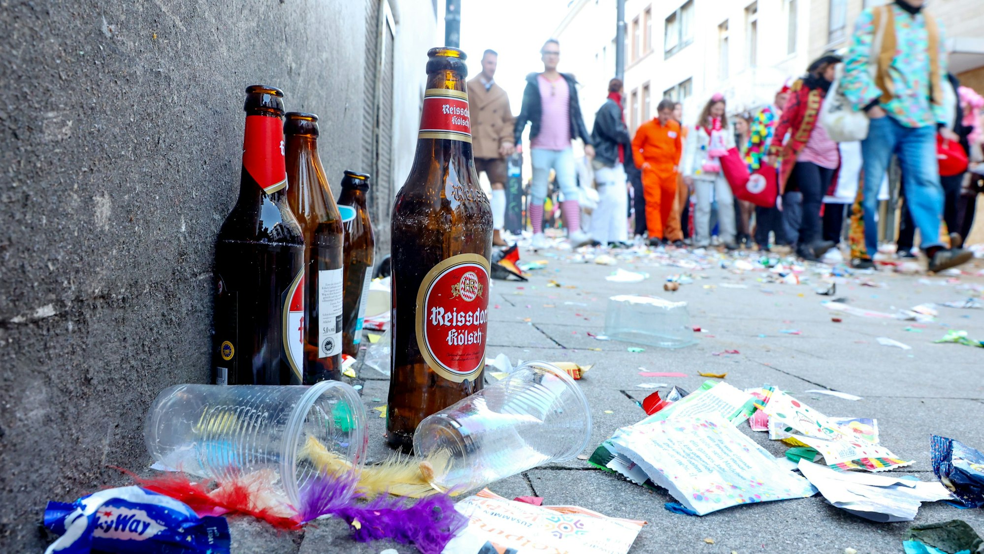 Zu sehen ist eine Ansammlung von Müll und leeren Bierflaschen auf einer Straße. Im Hintergrund sind Karnevalisten zu sehen.