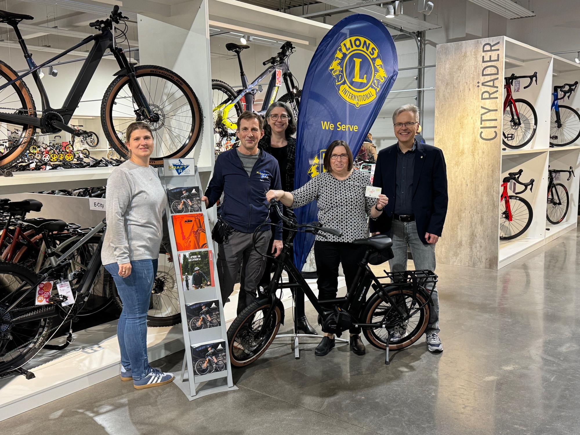 Auf dem Bild ist die Gewinnerin des E-Bikes mit mehreren Gratulanten und ihrem Gewinn im Fahrradgeschäft zu sehen.
