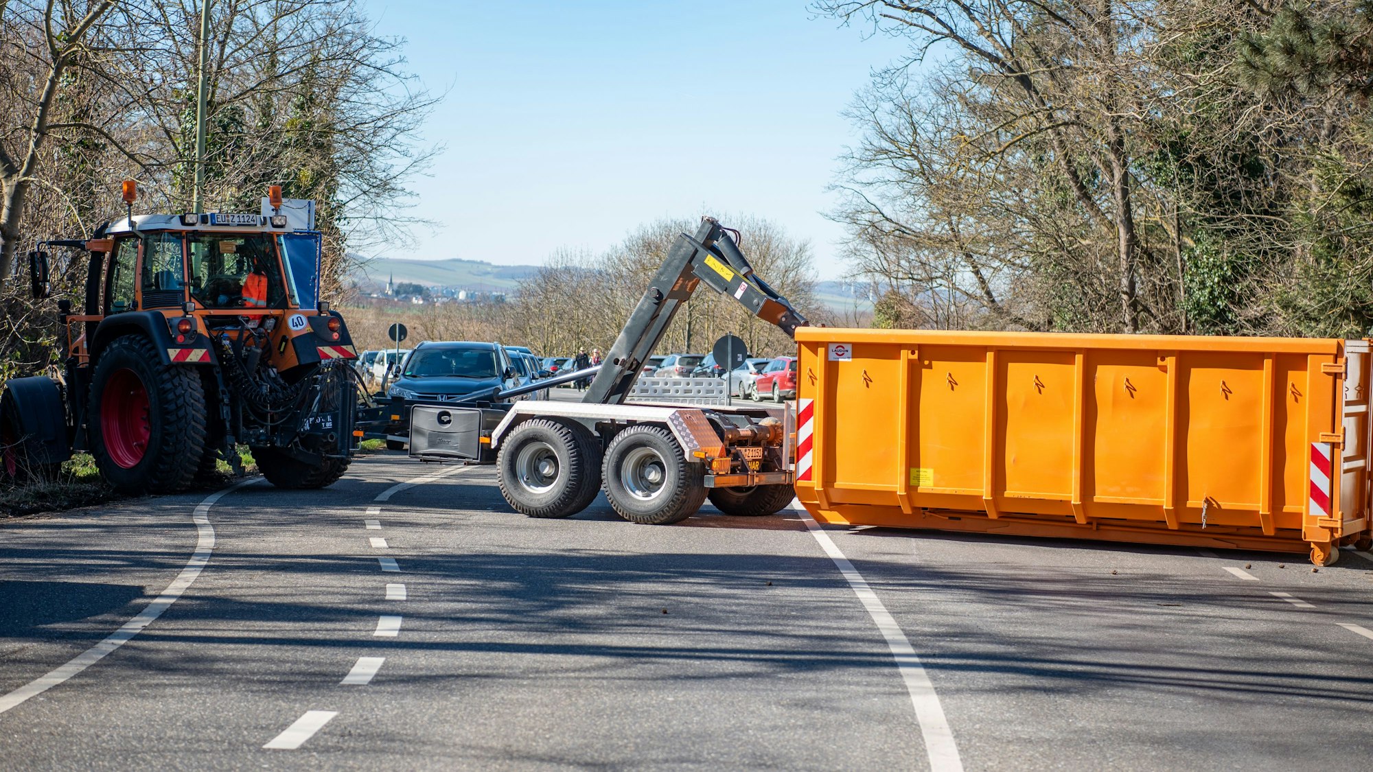 Ein Traktor mit Anhänger und ein oranger Container blockieren eine breite Straße.
