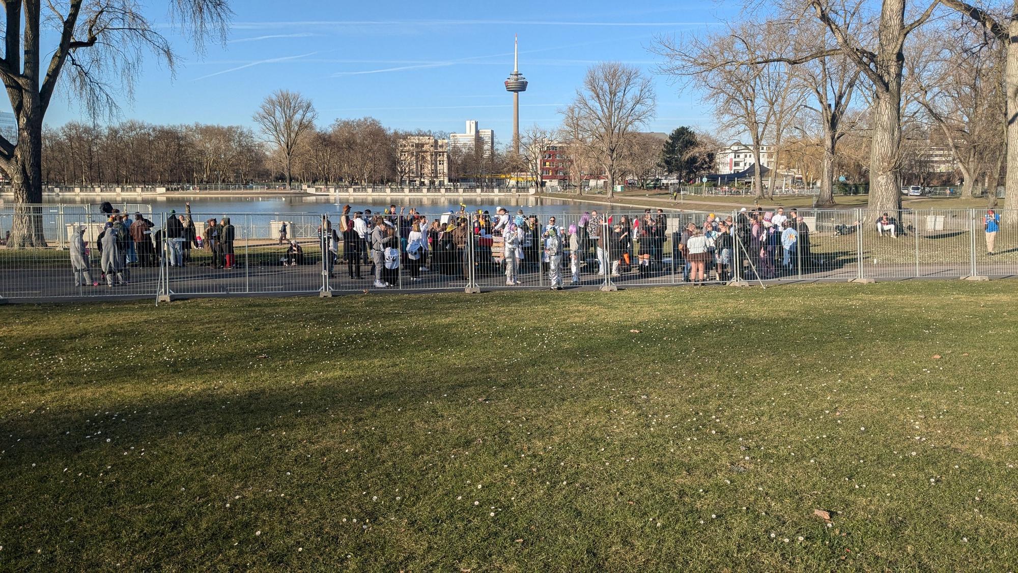 Eine Gruppe hat sich am Aachener Weiher mit einer Musikanlage versammelt und beschallt mit Popmusik eine Traube von Jecken
