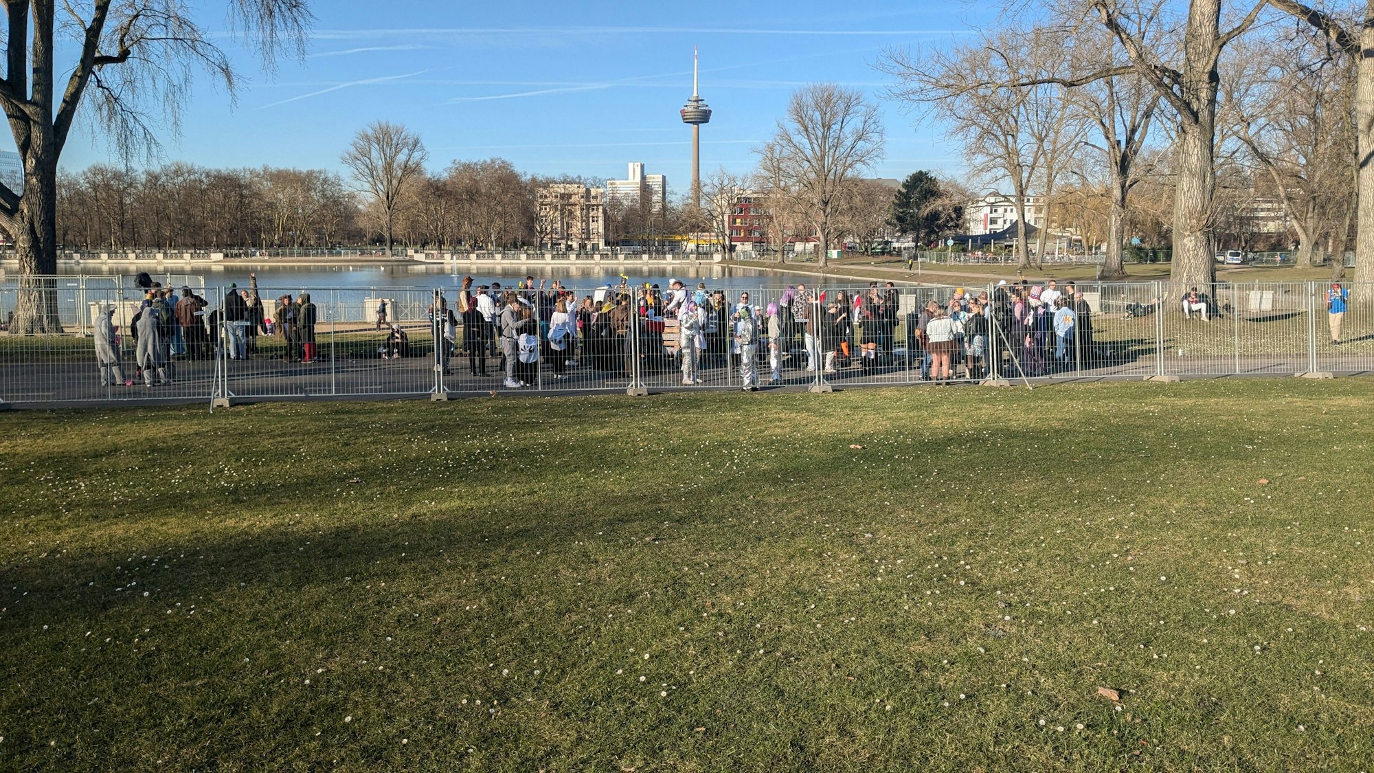 Eine Gruppe hat sich am Aachener Weiher mit einer Musikanlage versammelt und beschallt mit Popmusik eine Traube von Jecken