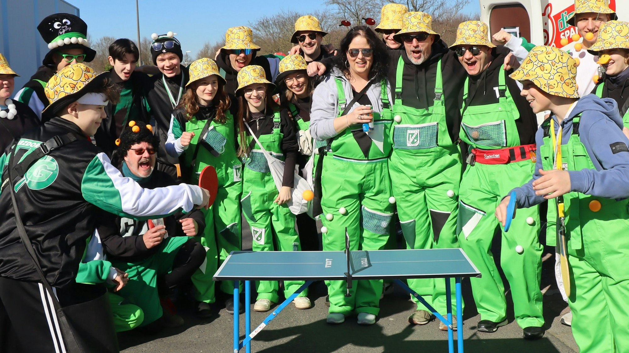 Auf dem Bild sind eine Gruppe in grünen Overalls und eine kleine Tischtennisplatte zu sehen, auf der zwei Jungen spielen.