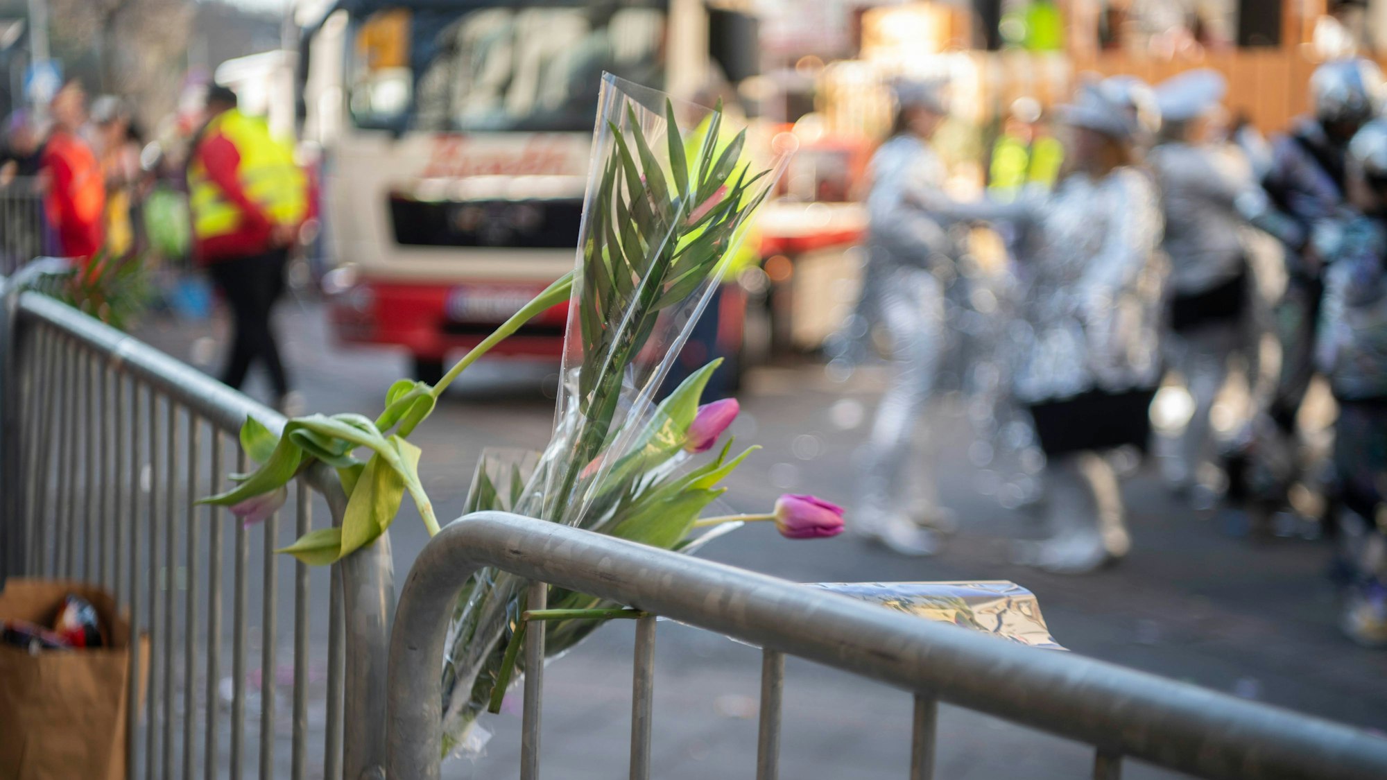 Im Rosenmontagszug in Euskirchen sind Tulpen-Sträußchen in ein Absperrgitter gesteckt. Im Hintergrund rollt ein Karnevalswagen an, verkleidete Jecke stehen auf der Straße.