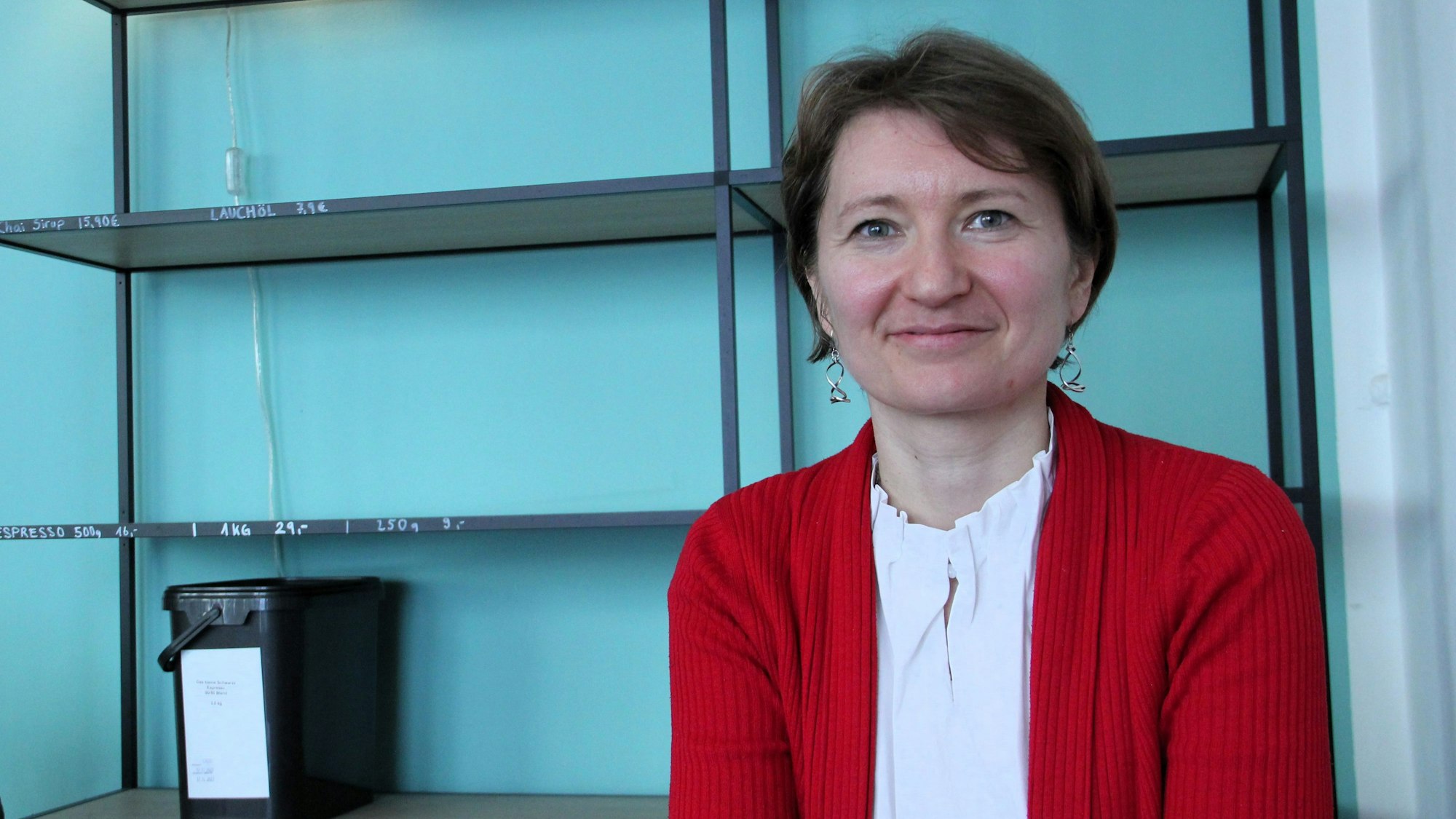 Eine Frau in weißer Bluse und roter Strickjacke sitzt vor einem Regal.