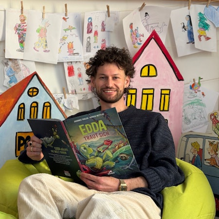 Ein Mann sitzt in einem Sitzsack und hält ein aufgeschlagenes Buch mit dem Titel „Edda taut sich“. Im Hintergrund hängen Kinderzeichnungen an einer Wand.