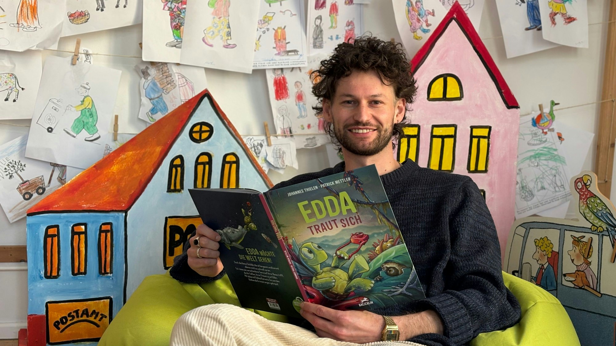 Ein Mann sitzt in einem Sitzsack und hält ein aufgeschlagenes Buch mit dem Titel „Edda taut sich“. Im Hintergrund hängen Kinderzeichnungen an einer Wand.