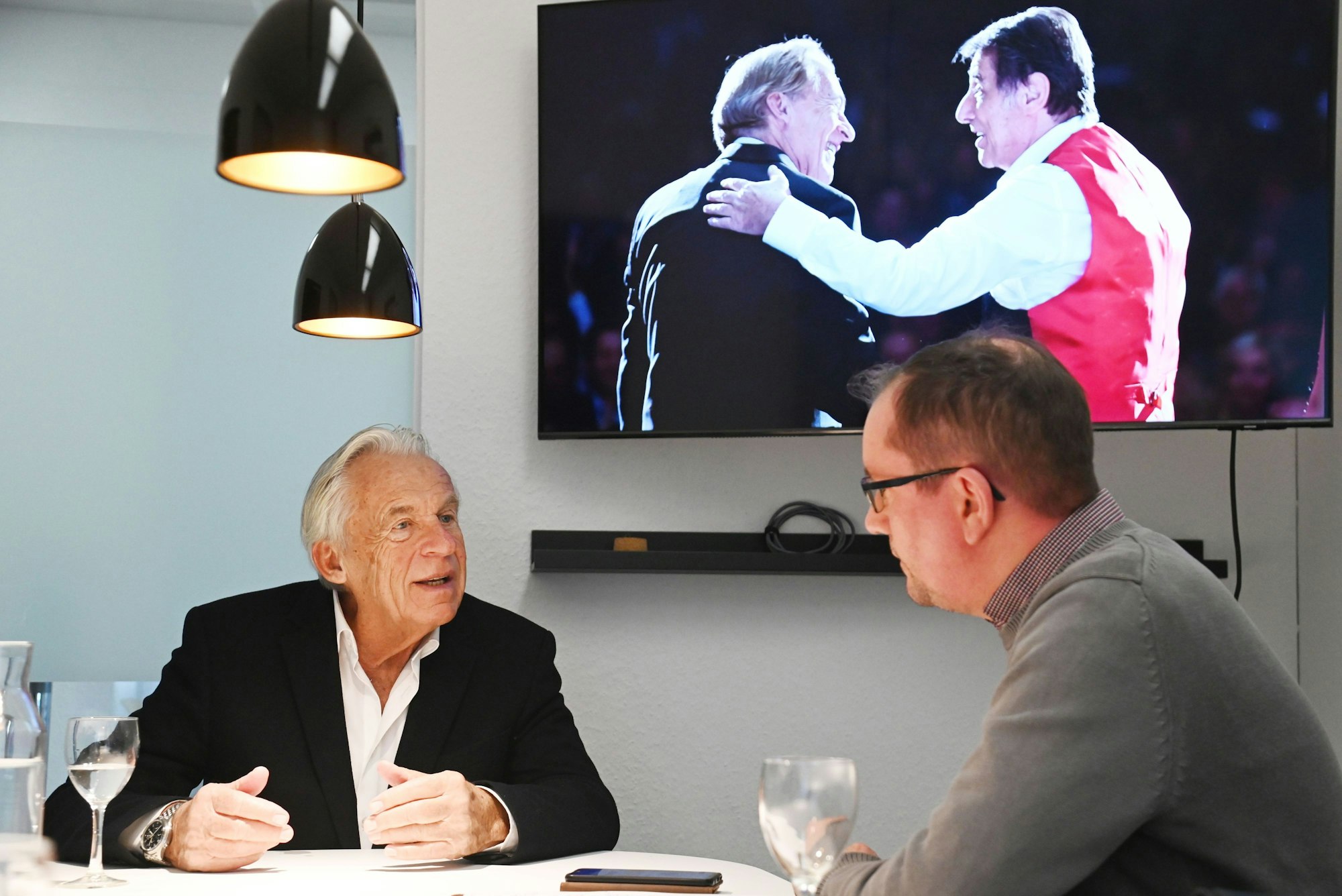 Pepe Lienhard beim Interview mit dem Kölner Stadt-Anzeiger in der Lanxess-Arena, im Hintergrund ist auf einem Bildschirm ein Foto von Lienhard und Jürgens aus dem Jahr 2014 zu sehen